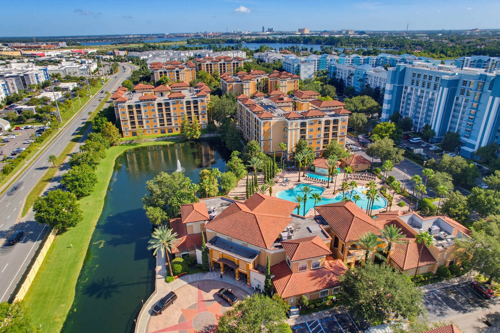 An aerial perspective showcasing a Mediterranean-style resort complex with terracotta roofs, landscaped grounds featuring a sparkling swimming pool, and a tranquil pond surrounded by palm trees and lush greenery.