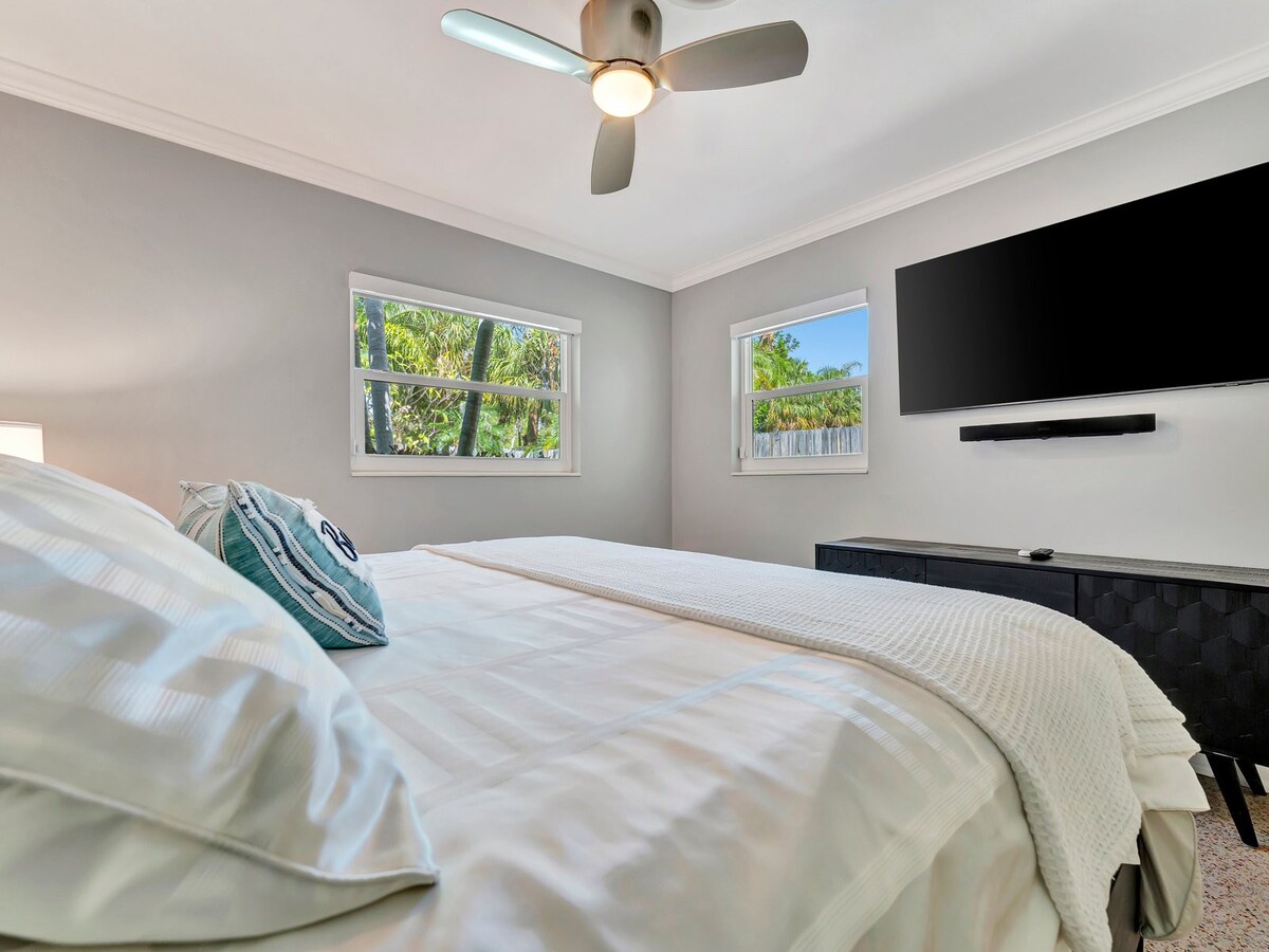 Unwind in your serene bedroom with tropical views framed by bright windows and modern touches like a ceiling fan and sleek entertainment setup.