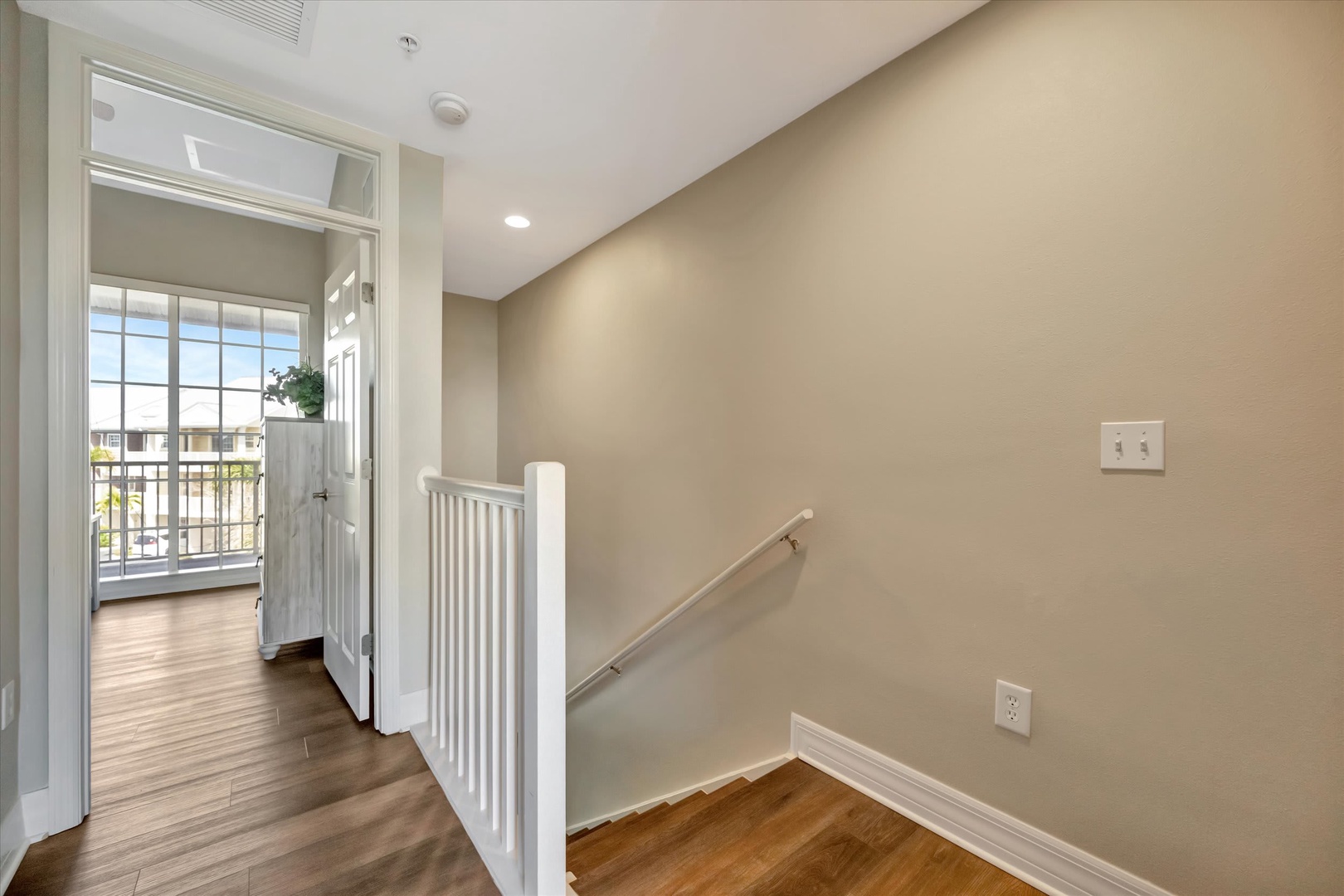 Step into your welcoming upstairs hallway, where warm hardwood floors and soft natural light create an inviting flow between rooms. Through the open doorway, catch a glimpse of the balcony that extends your living space outdoors.