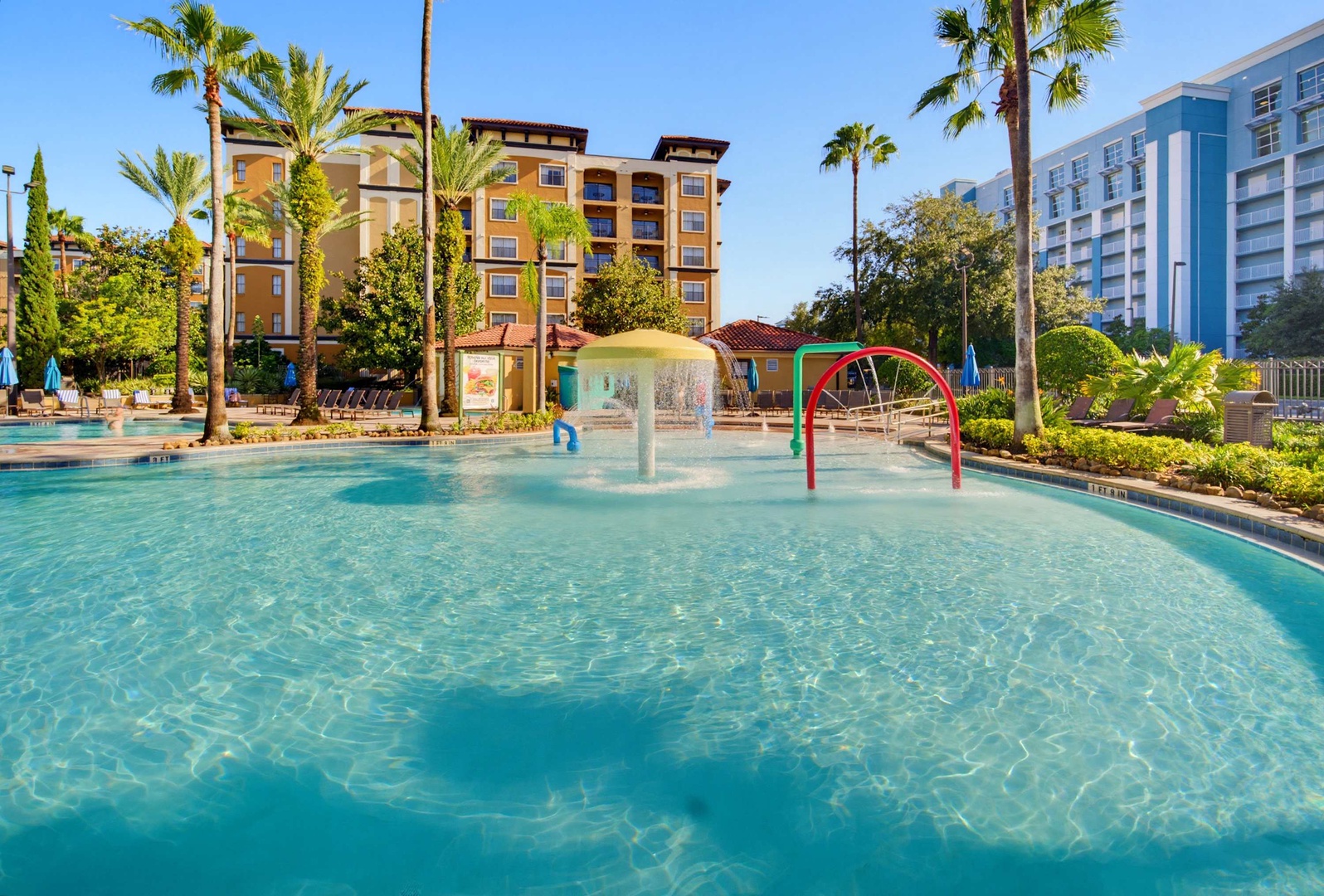 This family-friendly resort features a dedicated kids' splash pool with water fountains and play features, surrounded by lush tropical landscaping and palm trees under sunny Florida skies.