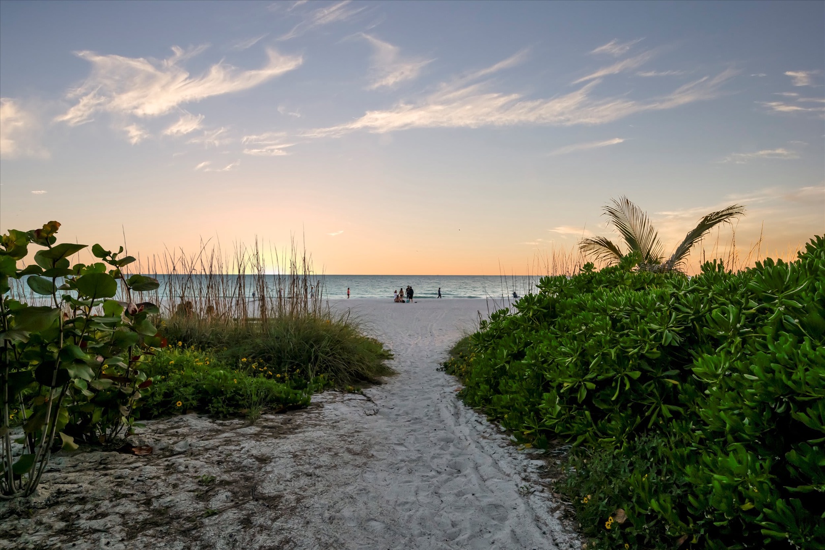 A sandy path winds through coastal vegetation toward a pristine white beach, where the turquoise water meets a pastel sky at sunset—a peaceful backdrop to the property's beachside location.