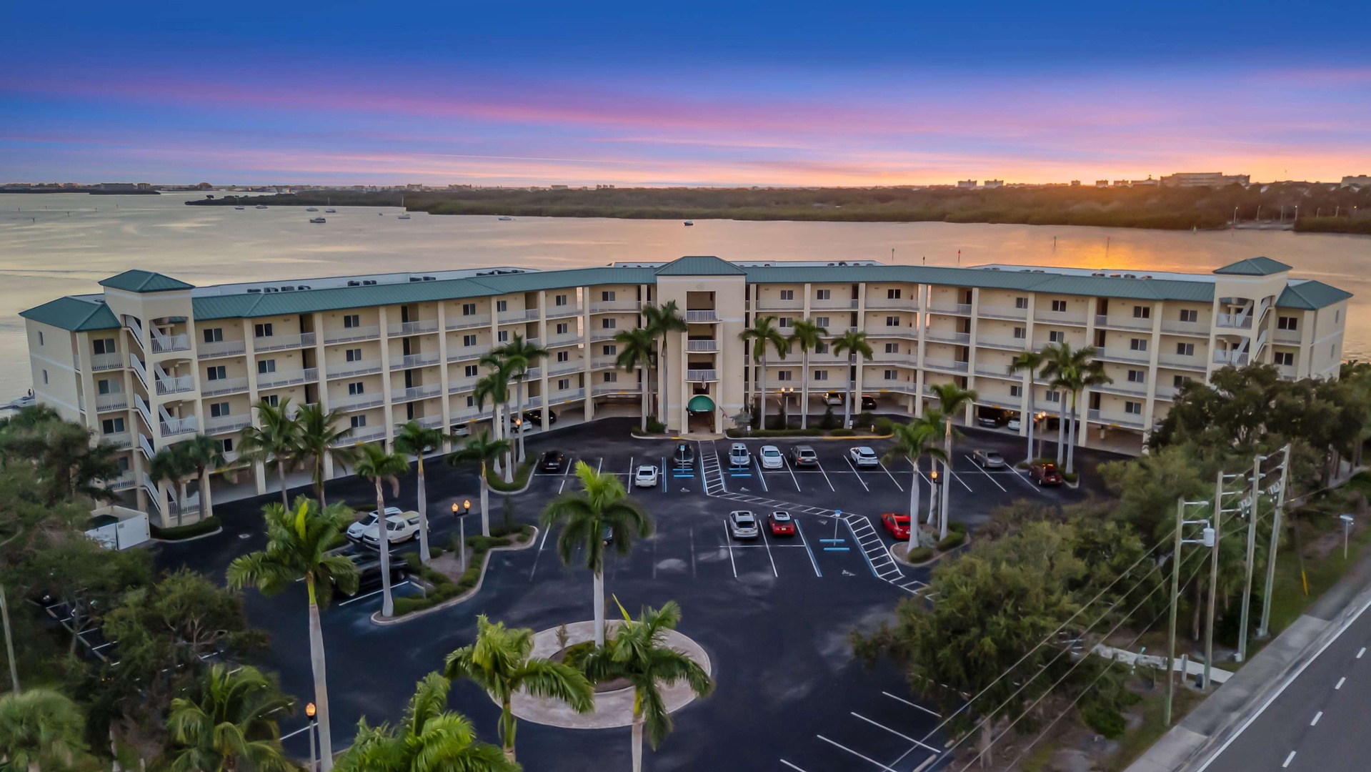 Aerial view of a waterfront vacation rental complex at sunset, showcasing tropical landscaping and guest parking near peaceful coastal surroundings.