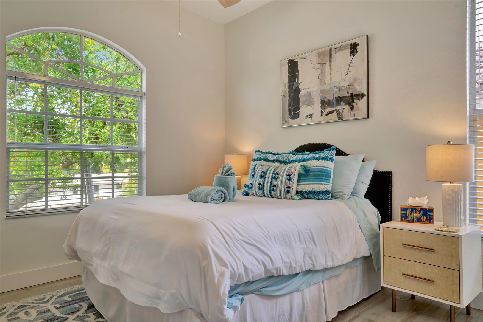 Step into your serene retreat where soft morning light filters through the arched window. Sink into crisp white linens adorned with turquoise accents and rest your head as gentle breezes and leafy views create your peaceful escape.