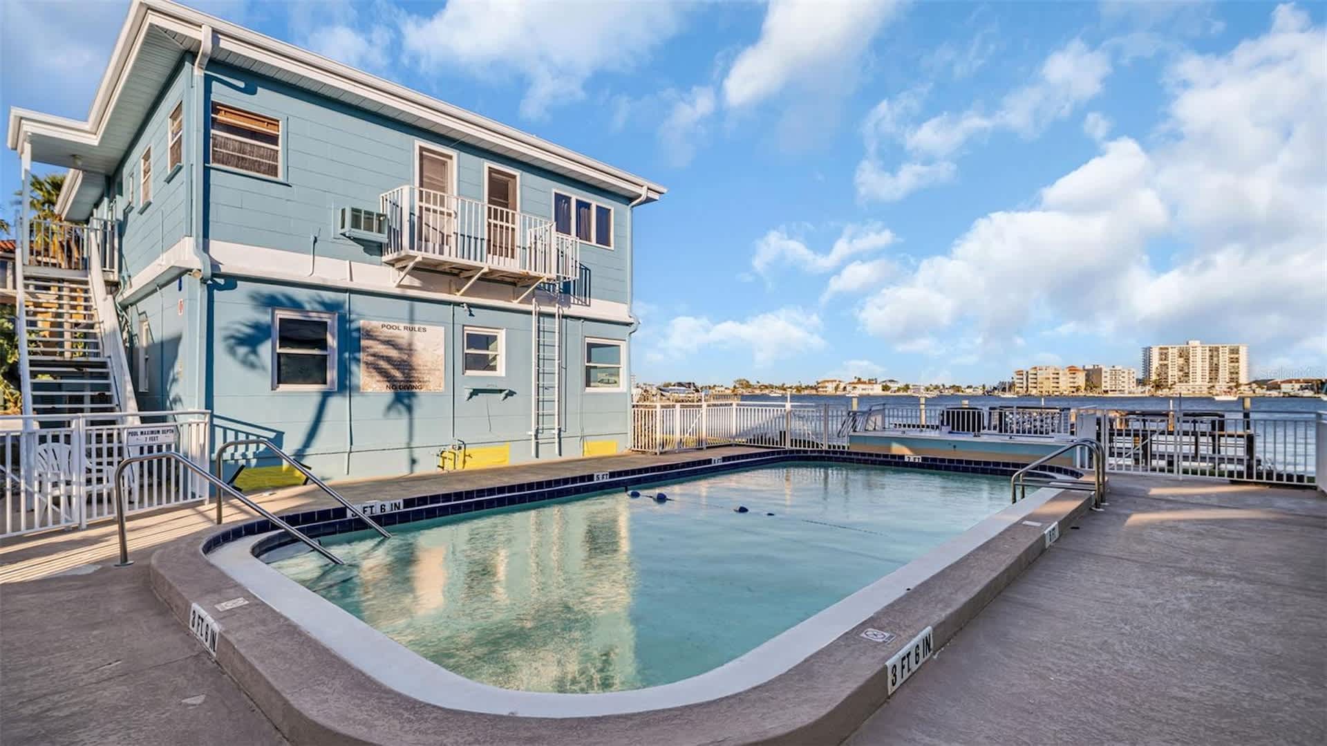 This waterfront property features a swimming pool overlooking calm bay waters, with nearby buildings visible across the shoreline. The two-story structure includes balcony access and outdoor decking in a quiet coastal setting.