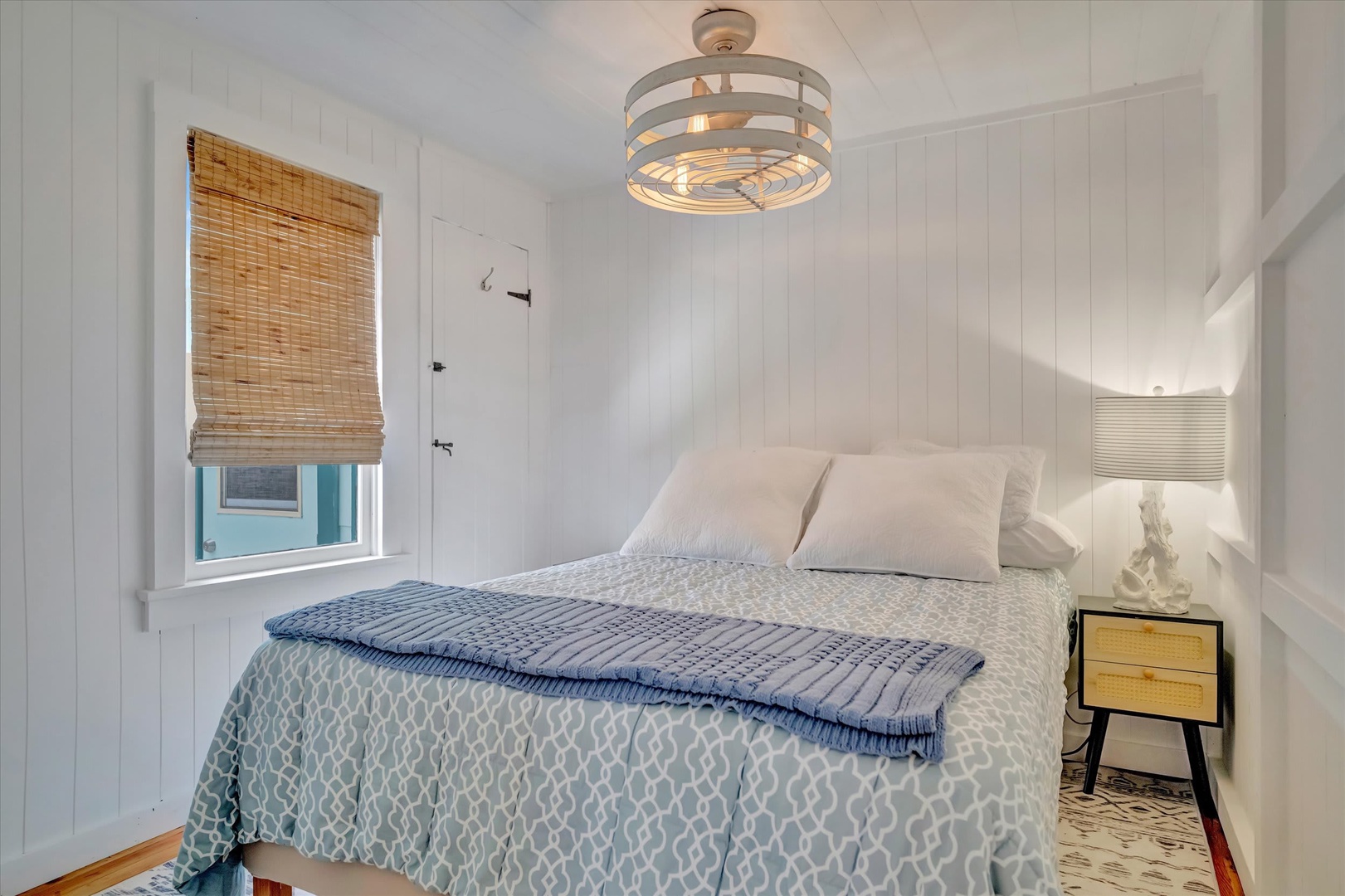 Slip into crisp white linens under a sculptural modern light fixture. This bright, coastal-style bedroom invites you to unwind in a serene space with charming wood paneling and natural bamboo accents.