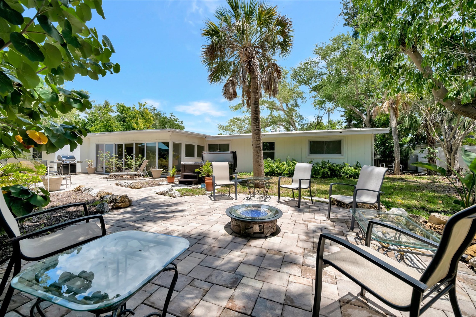 Spacious patio with cozy seating surrounds a central fire pit, perfect for evening gatherings under swaying palms. Your private backyard retreat awaits under sunny skies.