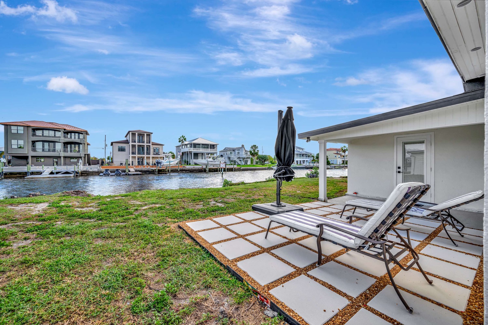 Waterfront property situated along a canal with boat docks and neighboring homes, featuring a covered patio with lounge chairs and grassy lawn area.