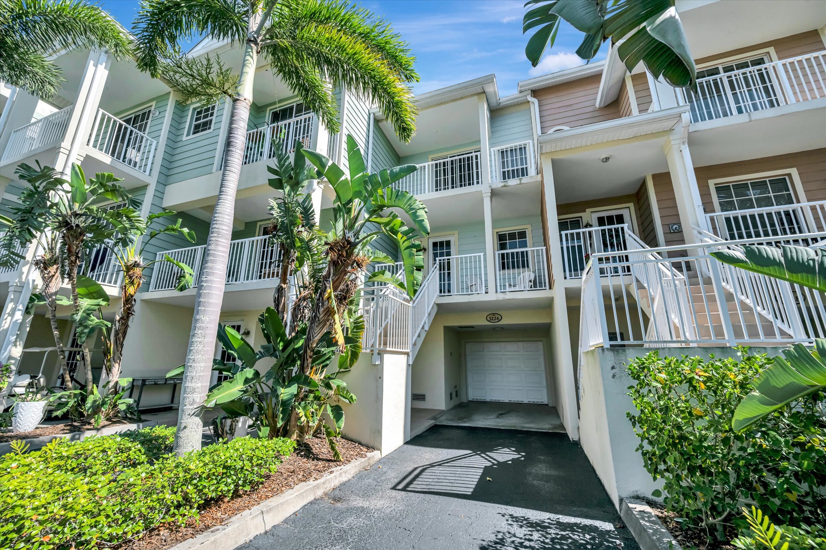 A coastal-style property building surrounded by lush tropical landscaping, featuring multiple levels of private balconies and ground-floor parking access beneath swaying palms.