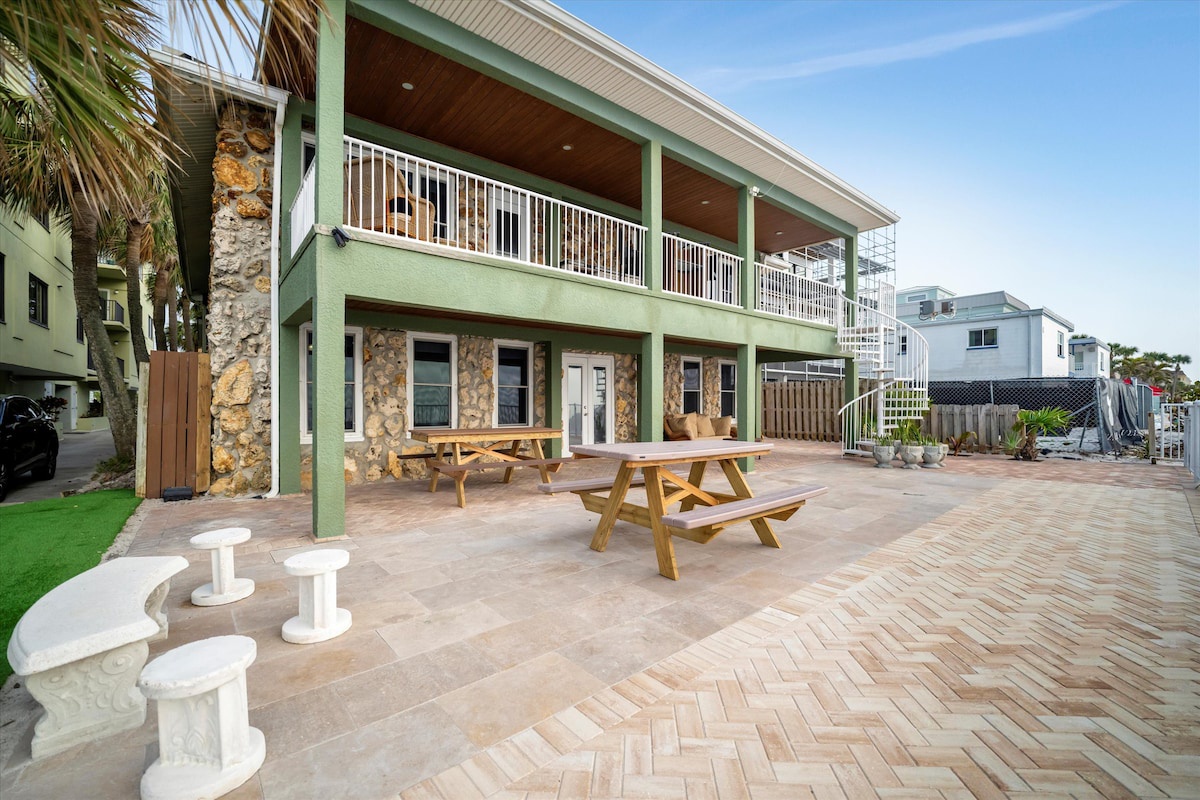 A coastal property featuring picnic tables, tropical palms, and herringbone brick patio—an inviting outdoor space near other beach neighborhood buildings.