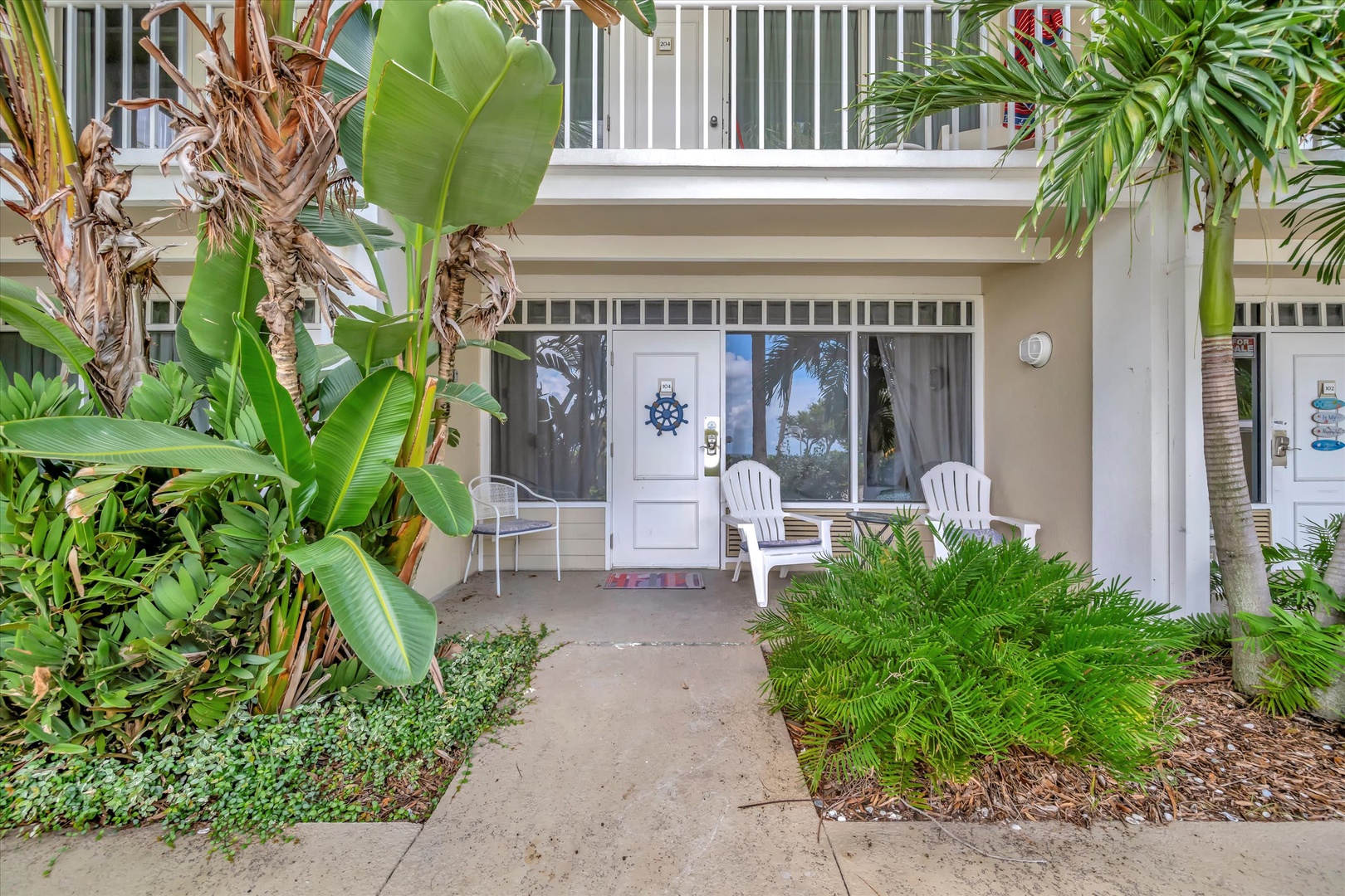 Tropical landscaping frames this welcoming ground-floor entrance with private patio and second-floor balcony overlooking lush greenery, enhanced by nautical charm and Adirondack chairs.