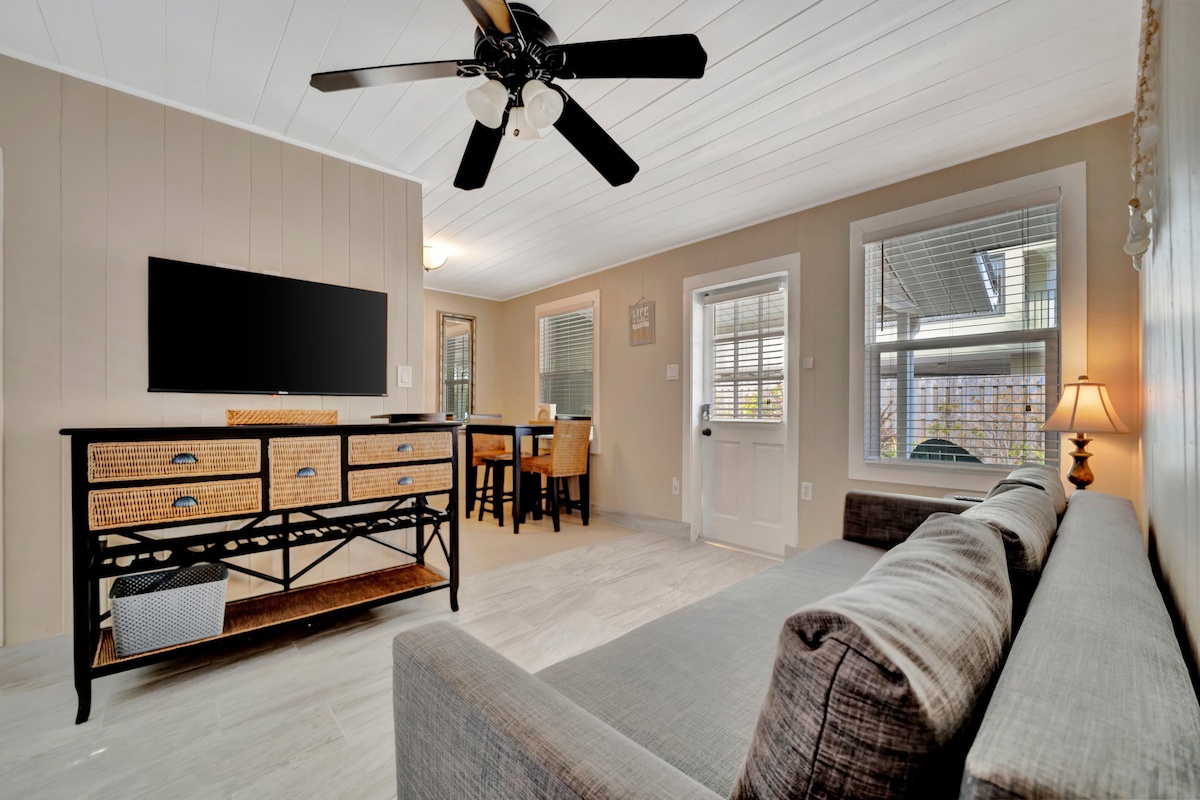 Relax in your bright, open living space with comfortable seating, a ceiling fan, and natural light streaming through large windows—perfect for unwinding after your beach adventures.