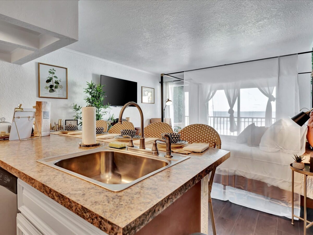Bright coastal kitchen flowing into living and dining areas, featuring a shimmering copper sink and billowing curtains that frame ocean breezes in this serene beach retreat.