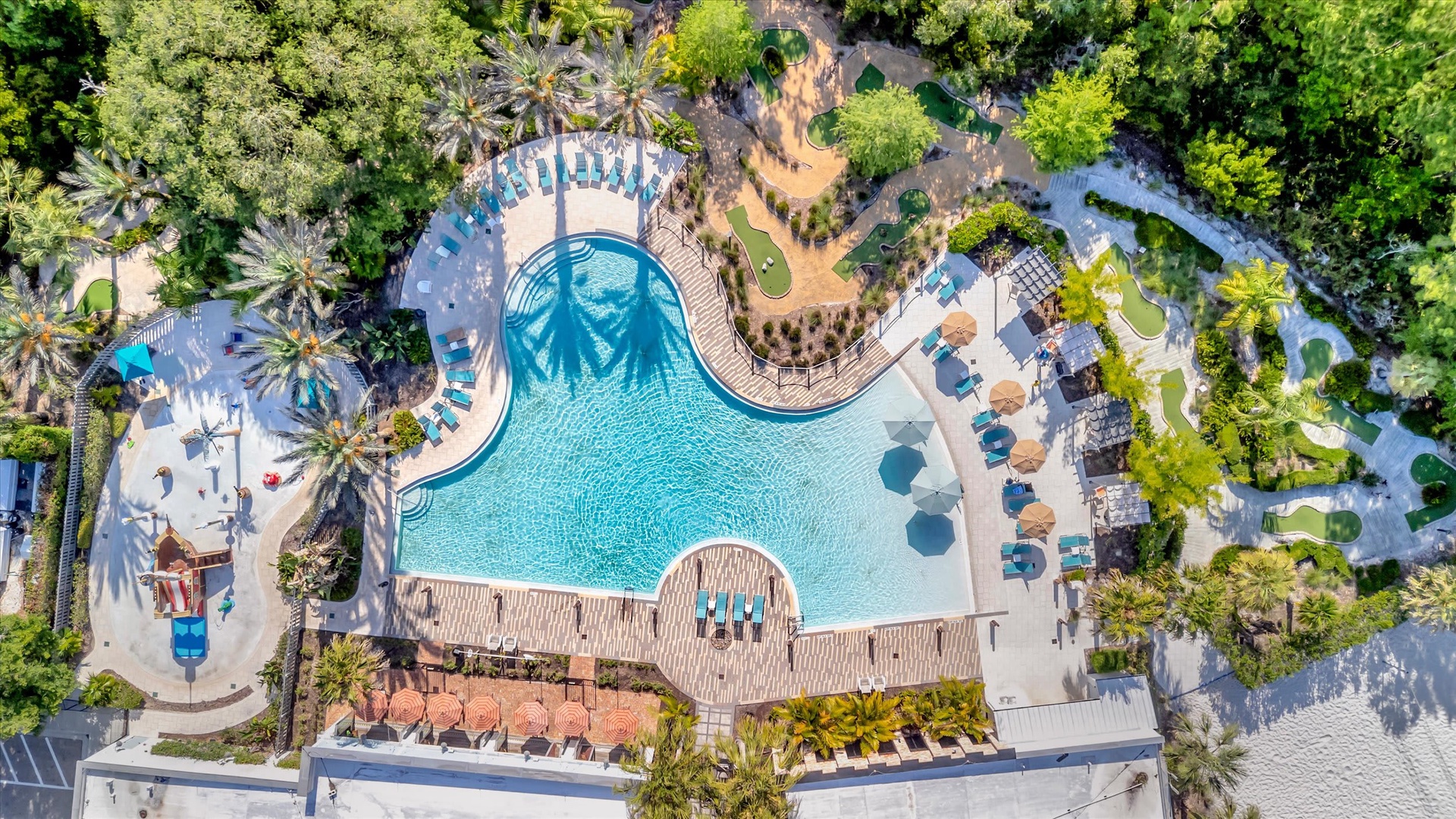 An aerial view of the resort-style pool area, surrounded by lush landscaping, palm trees, and ample deck space with lounge chairs and umbrellas perfect for sunny days.