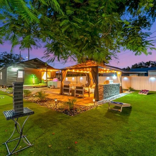 Twilight transforms the backyard into a dreamy escape—string lights glow above the pergola, casting a warm shimmer over the dining and lounge area nestled in lush green grass beneath swaying palms.