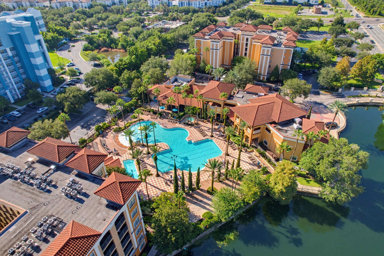 An aerial view reveals the resort-style property with Mediterranean architecture, featuring a sprawling pool complex surrounded by palm trees, lush landscaping, and a scenic lake.
