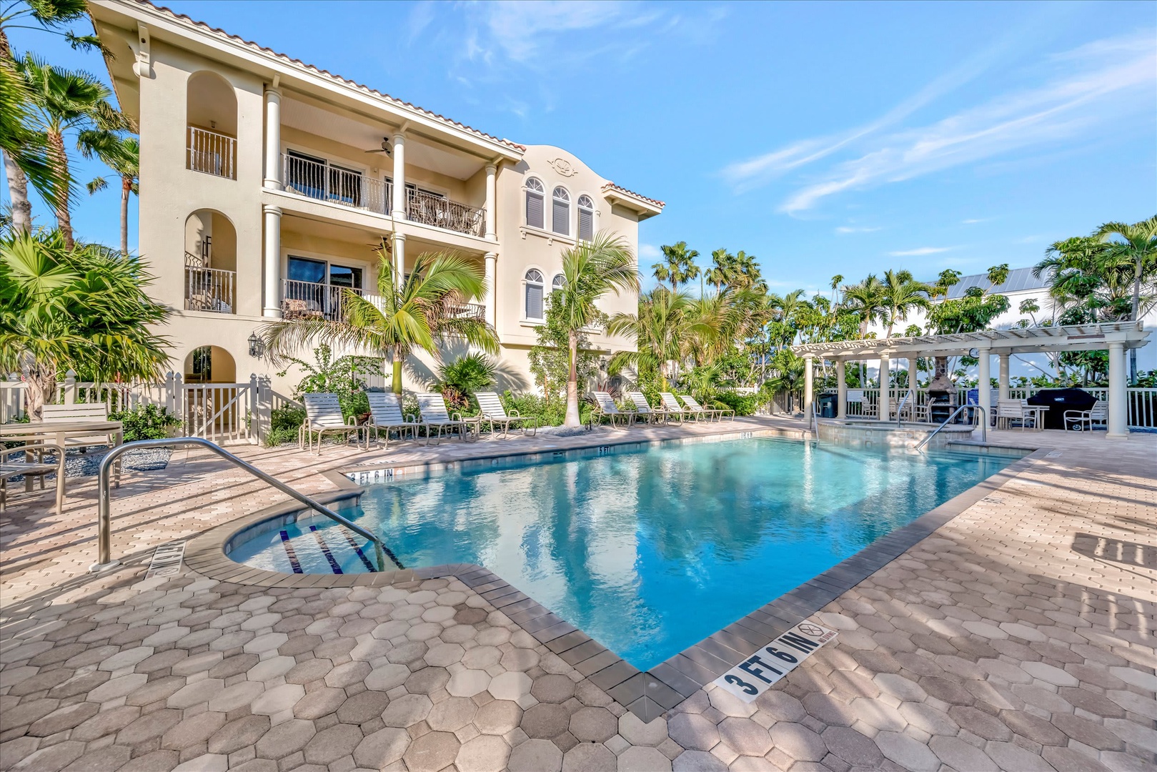 A sparkling pool invites you to cool off under the tropical sun, surrounded by lush palms, sunloungers, and elegant Mediterranean-style architecture with shaded seating areas.
