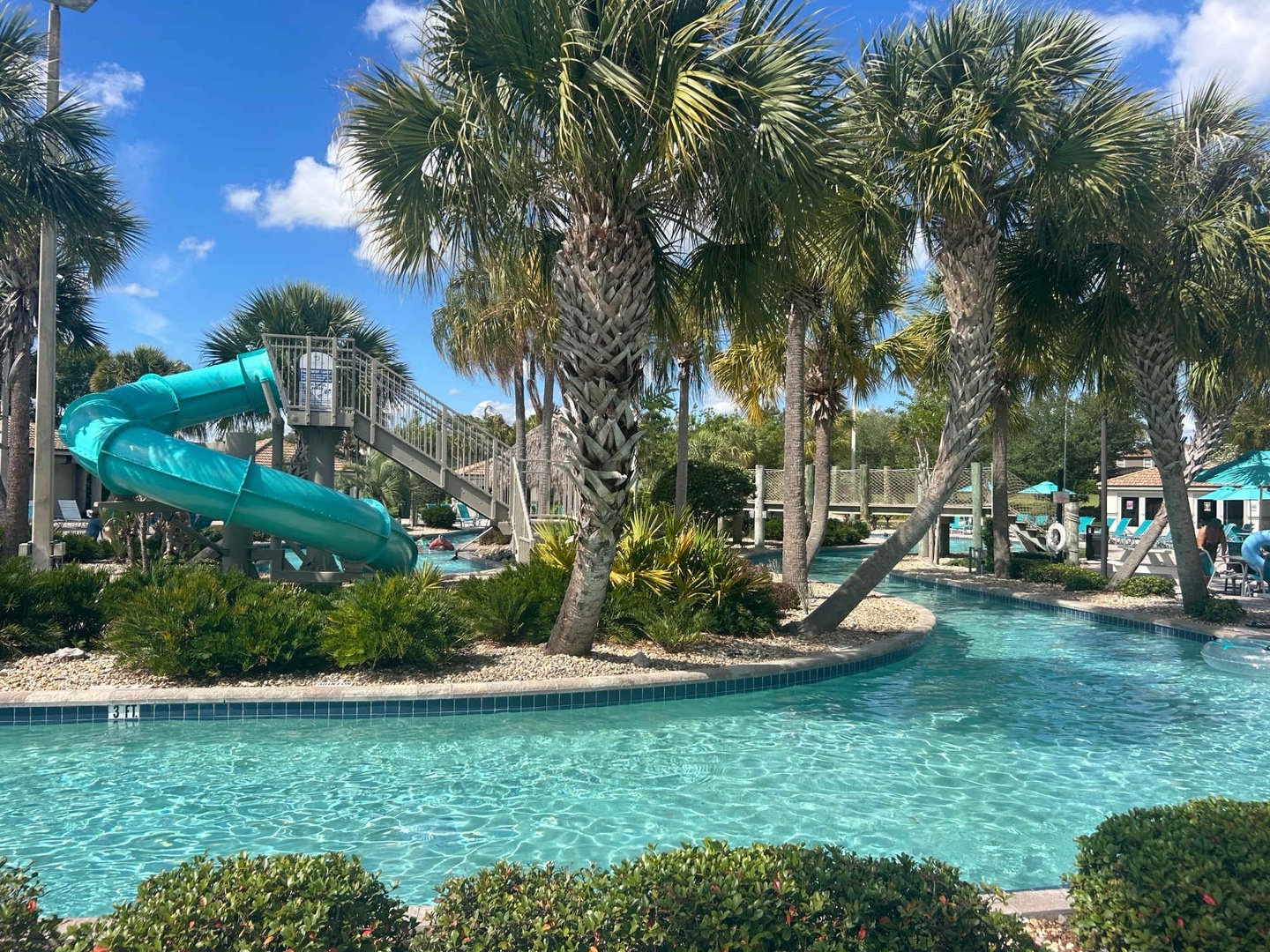 A sparkling aqua pool winds through swaying palms, with a thrilling water slide beckoning adventure under sunny skies. Your tropical escape awaits in this lush, vibrant oasis.