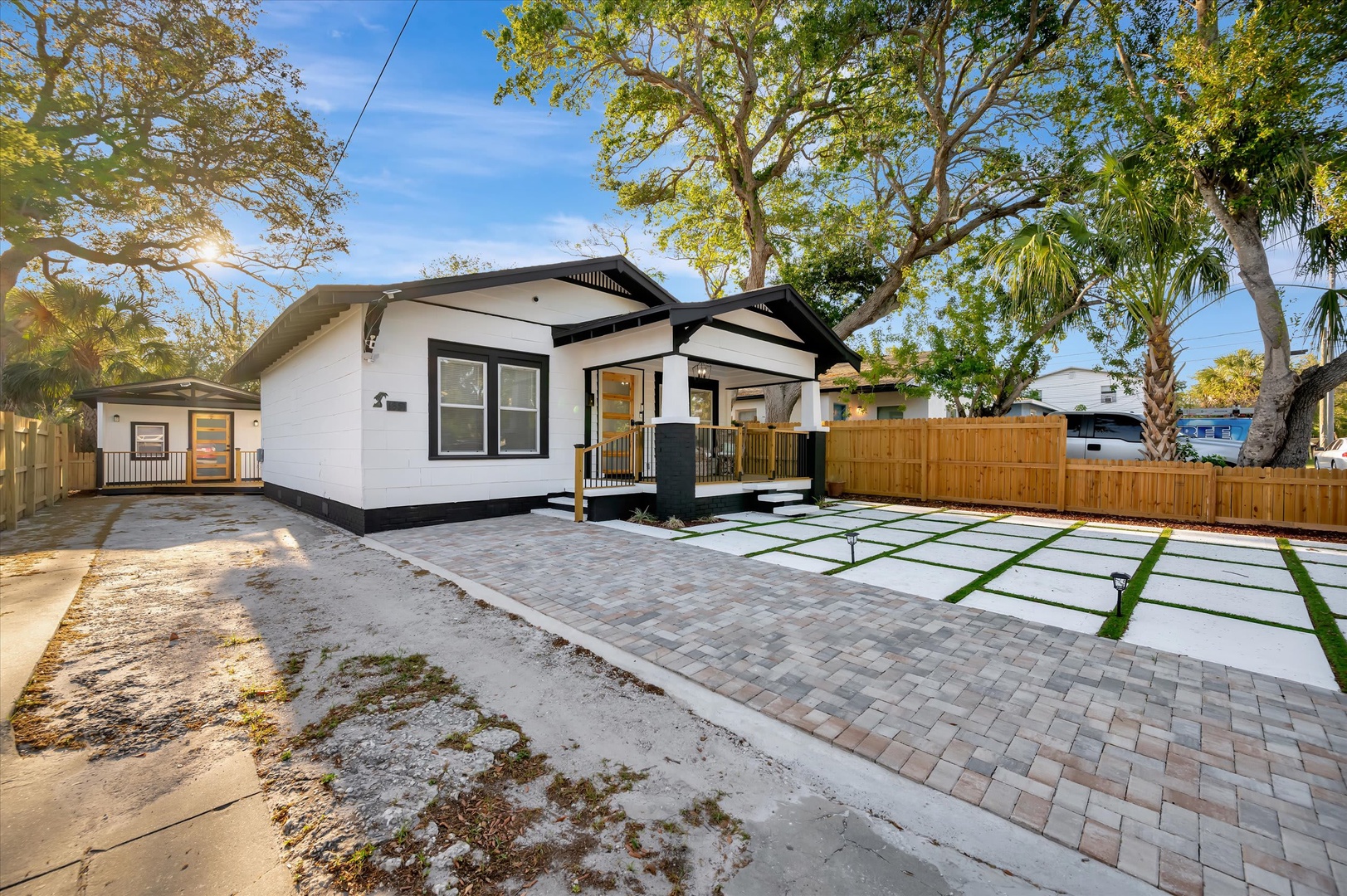 Modern vacation home featuring a stylish white exterior with black trim, surrounded by mature trees and a spacious driveway with paver stones and grass strips.