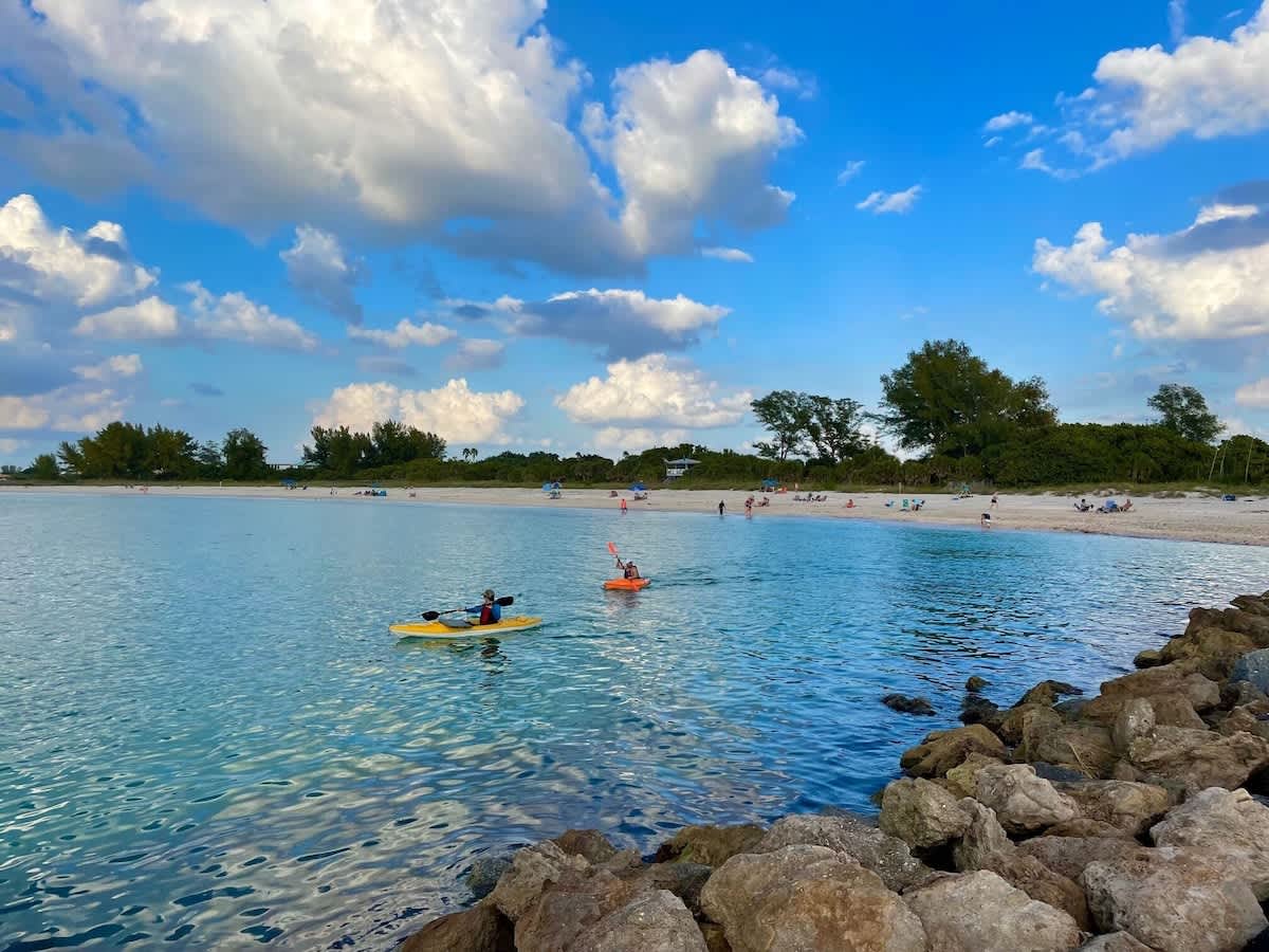 A scenic waterfront area near the property features calm turquoise waters perfect for kayaking, with a sandy beach and lush tree-lined shore beneath a brilliant summer sky.