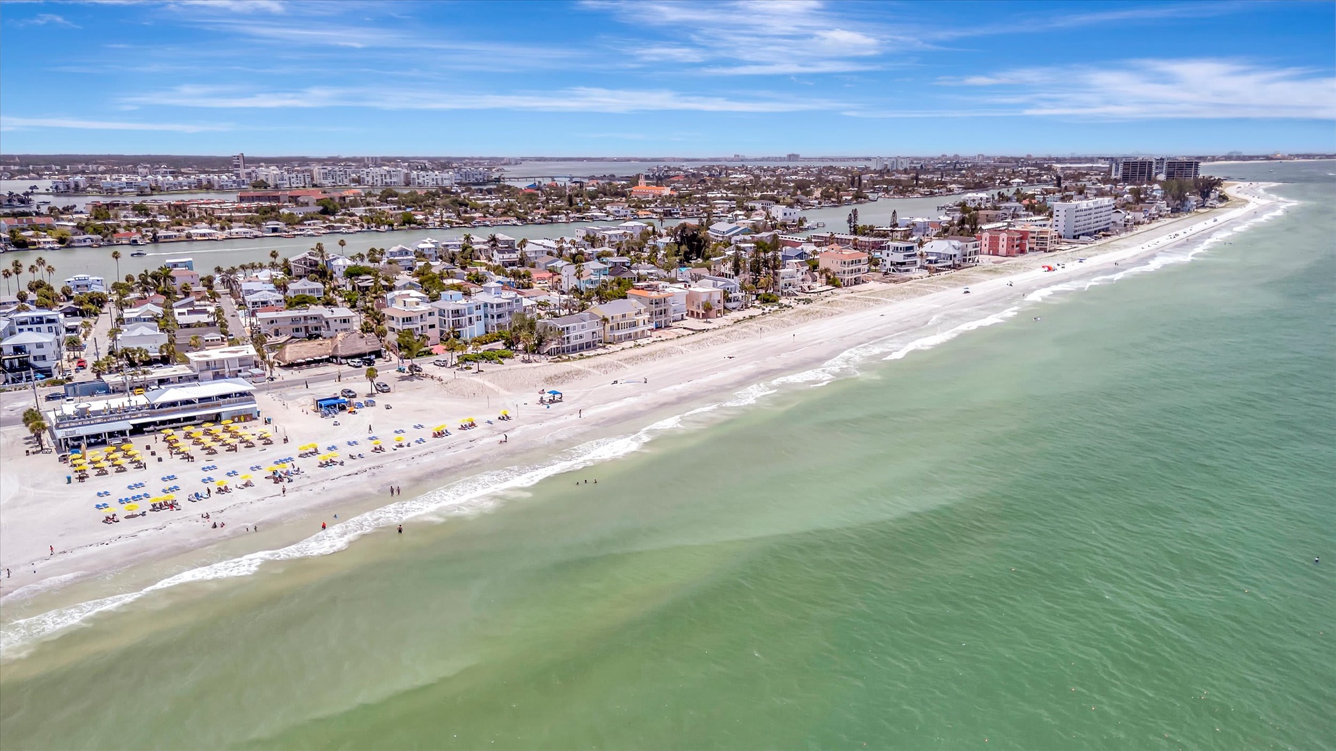An aerial view captures the property's beachfront location along pristine sandy shores and calm turquoise waters, with palm trees and coastal buildings dotting the surrounding neighborhood.
