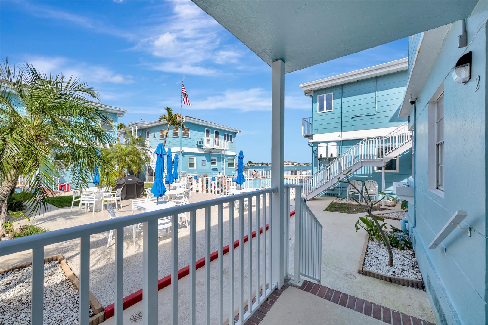 A charming waterfront property featuring classic coastal architecture with turquoise-painted buildings, palm trees, and access to a peaceful beach area with lounge chairs and umbrellas.