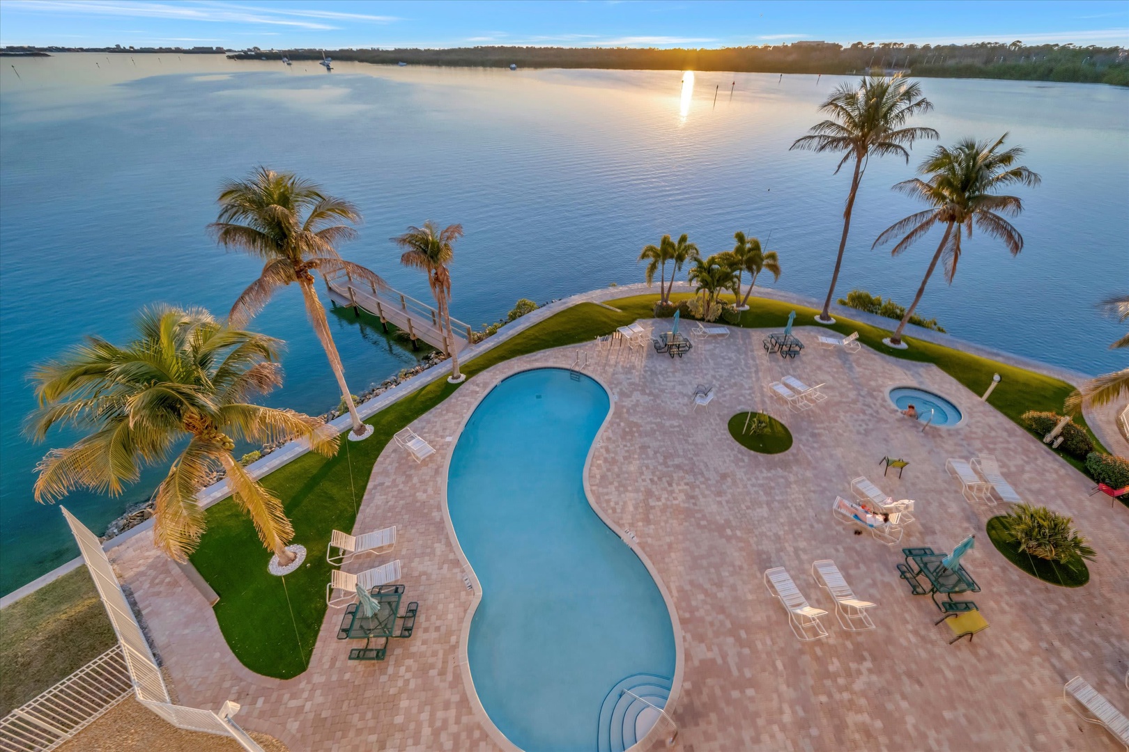 An aerial view captures the waterfront property at sunset, featuring a winding pool, hot tub, and private dock extending into serene waters surrounded by tropical palms.