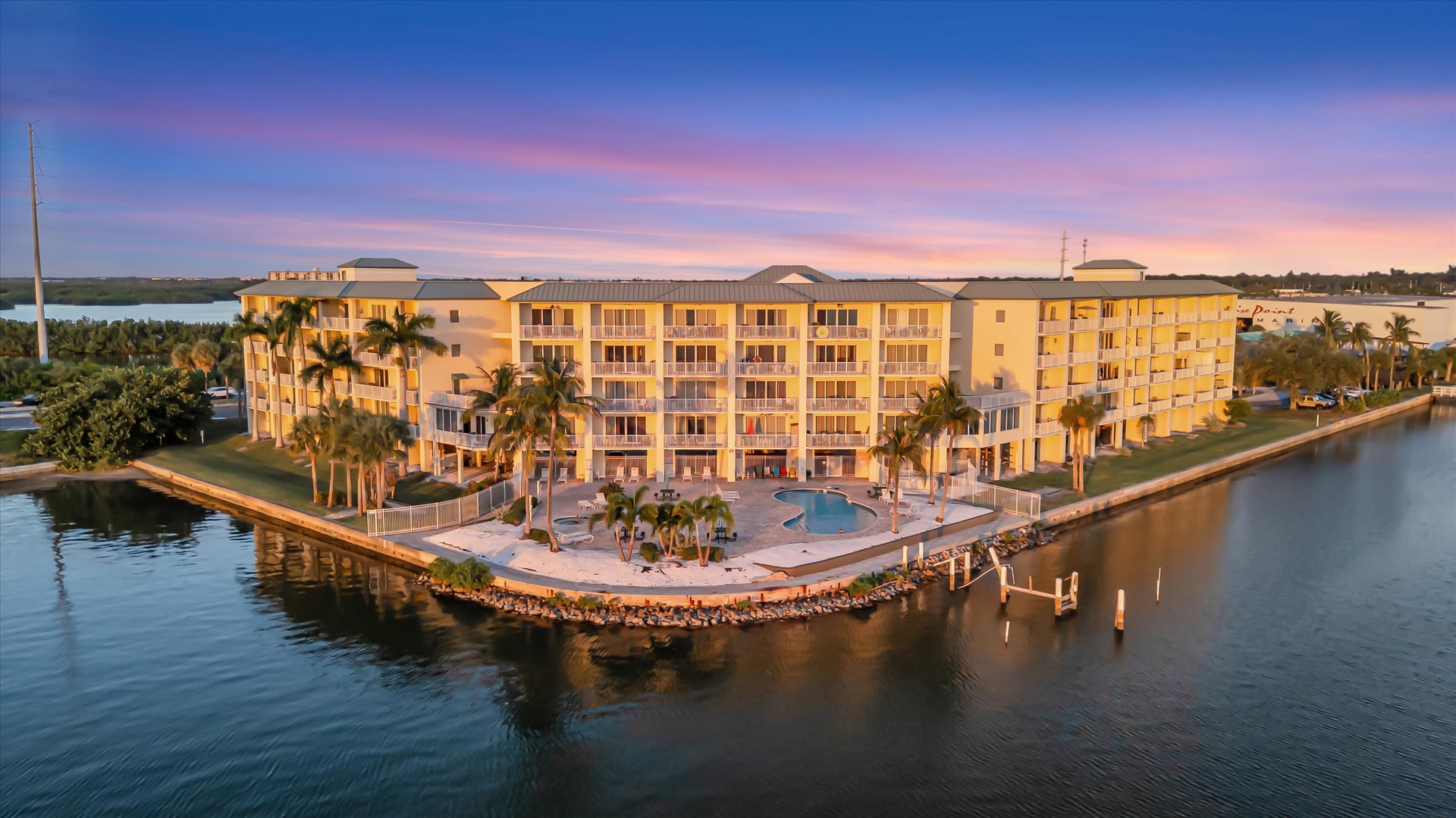 The resort complex sits on waterfront property, with multiple buildings, palm trees, a pool, and a private dock. Captured during a vibrant sunset, the property is surrounded by calm water and accessible by boat.