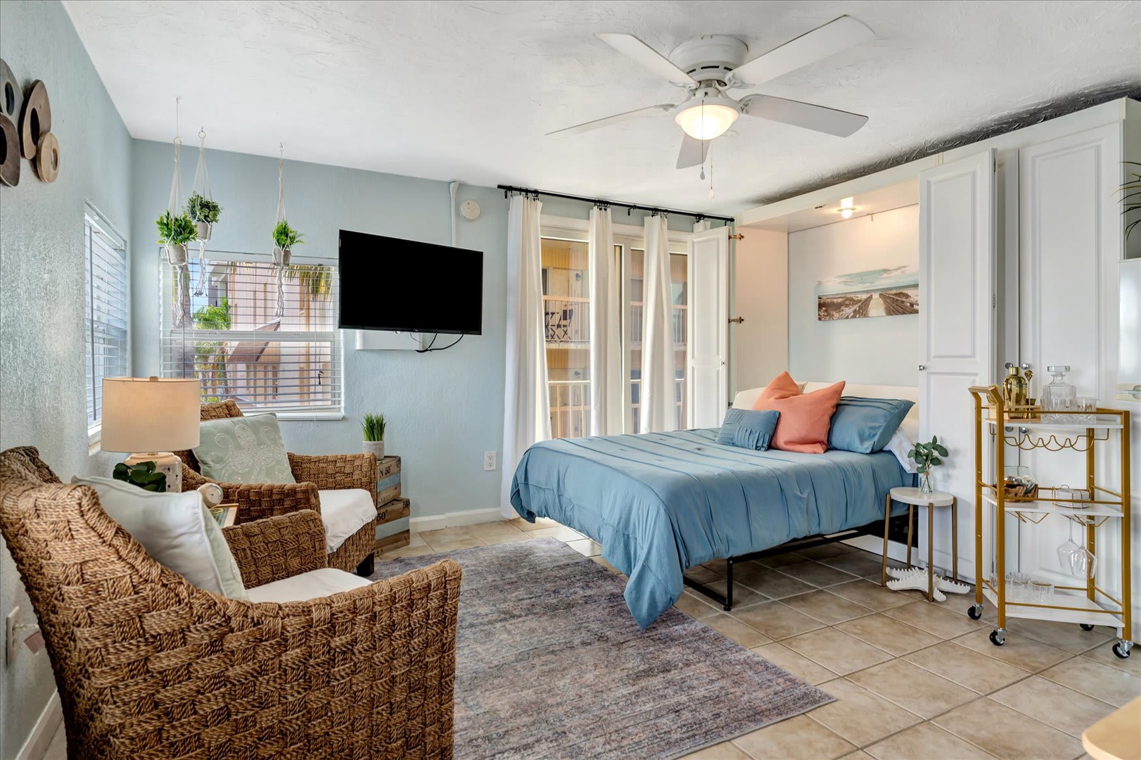 Relax in your coastal-inspired studio with soft blue tones, wicker seating, and a cozy bed. Natural light flows through the window while you unwind under the ceiling fan after a day exploring the area.