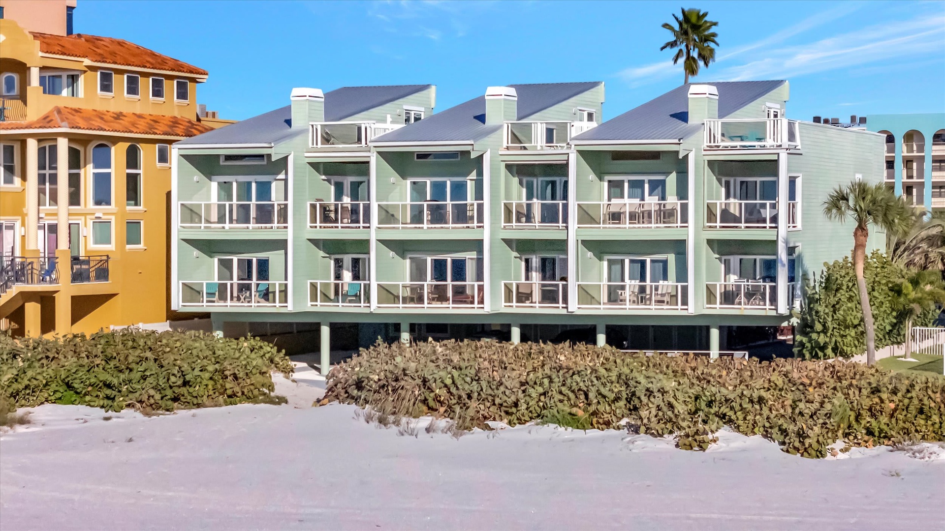 A beachfront vacation rental building stands directly on sandy shores, surrounded by coastal vegetation and neighboring properties, offering direct beach access for guests.