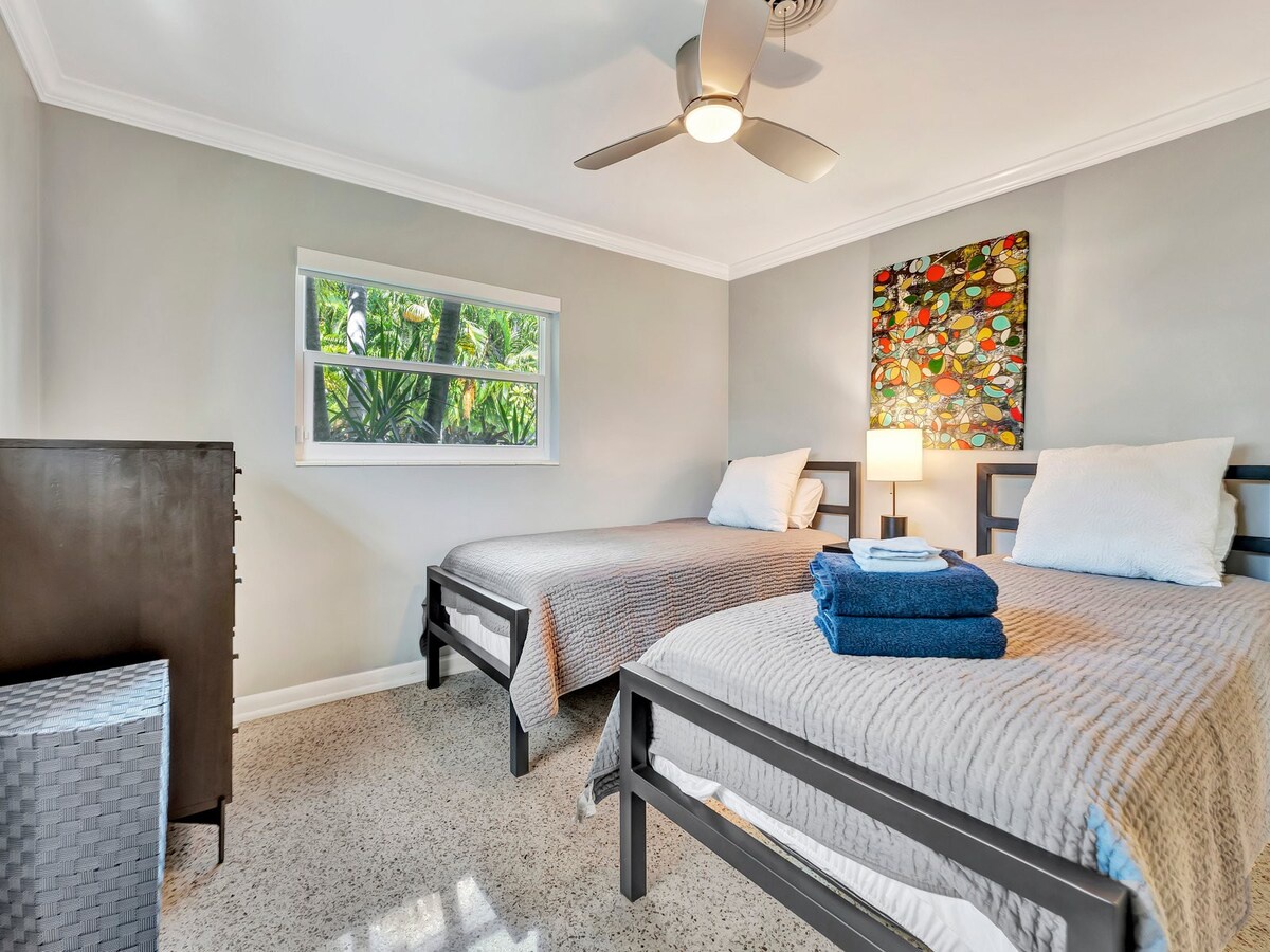 Your serene guest bedroom features two plush beds dressed in soft linens, a ceiling fan for comfort, and a window framing lush tropical greenery—perfect for restful nights and peaceful mornings.