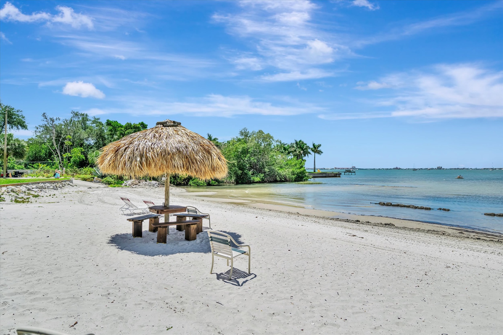 A private beach area featuring a thatched palapa and seating where the calm bay waters meet pristine sand, surrounded by lush tropical greenery and open skies.