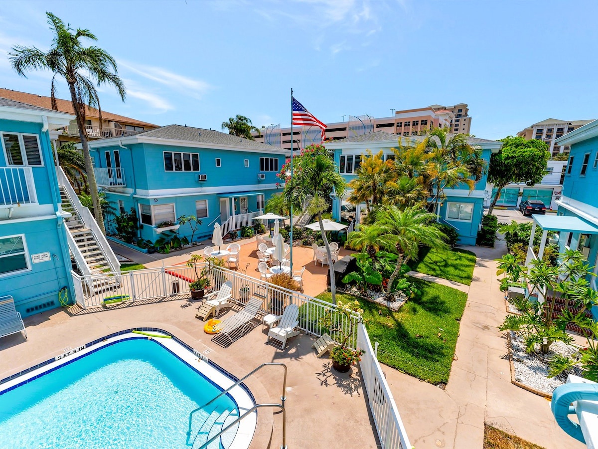 A vacation rental complex with a central courtyard featuring a swimming pool, landscaped gardens, and lounge areas. Surrounded by bright blue residential buildings, palm trees, and nearby urban buildings visible in the background.