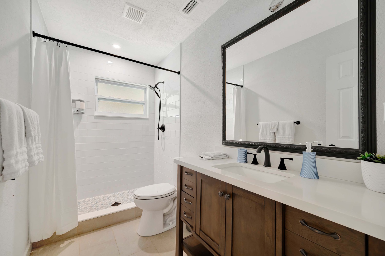 Your private bathroom features a spacious walk-in shower, modern vanity with crisp white countertops, and thoughtful touches like fresh towels and natural light from the window.