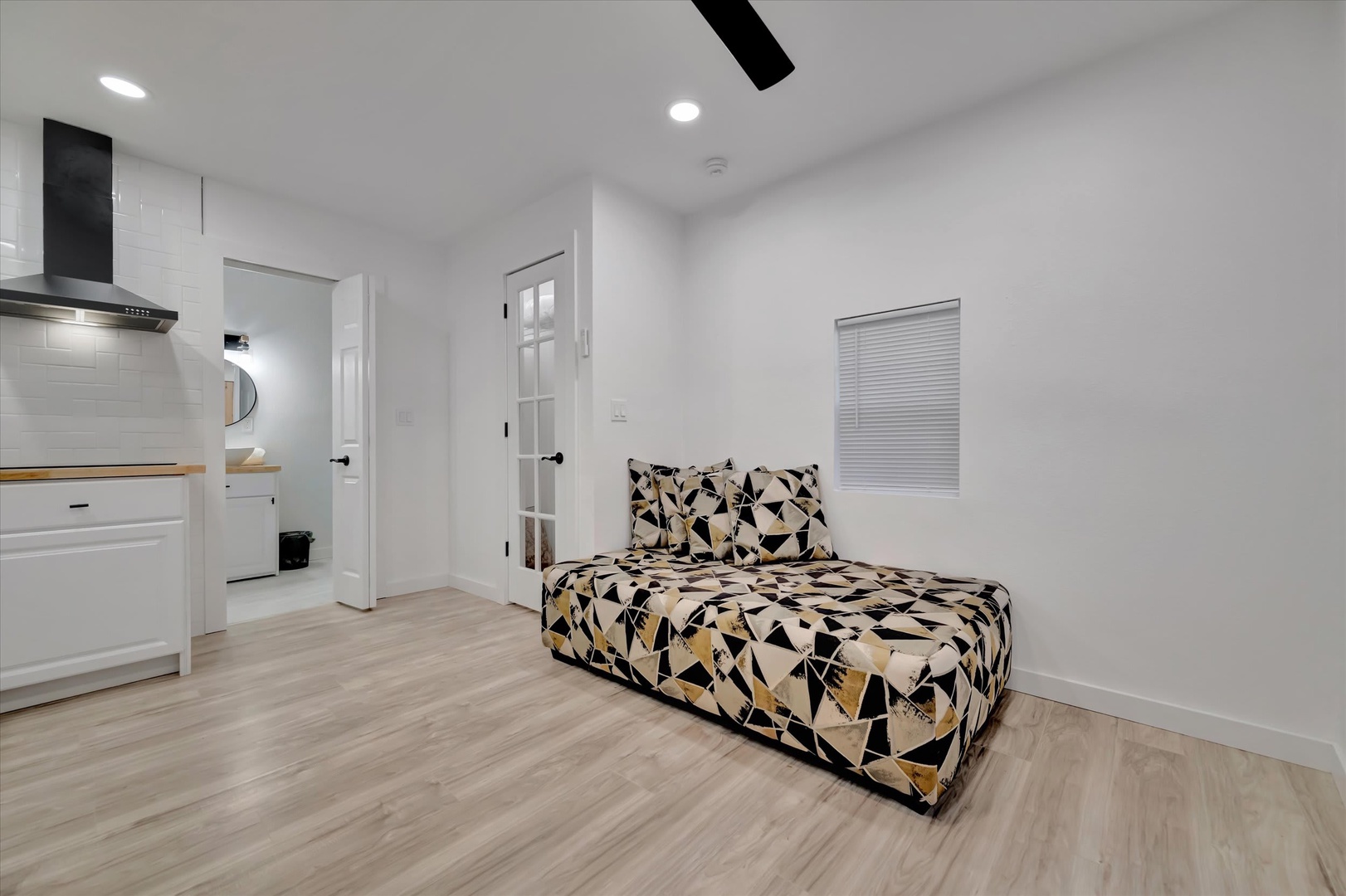Your modern studio retreat features sleek wood-style flooring and a fresh, minimalist aesthetic. Relax on the cozy daybed while enjoying natural light through the window, with a compact kitchenette and private bathroom just steps away.