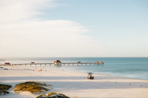 A pristine beachfront setting awaits, with soft white sands stretching toward calm turquoise waters and a historic pier reaching into the horizon.
