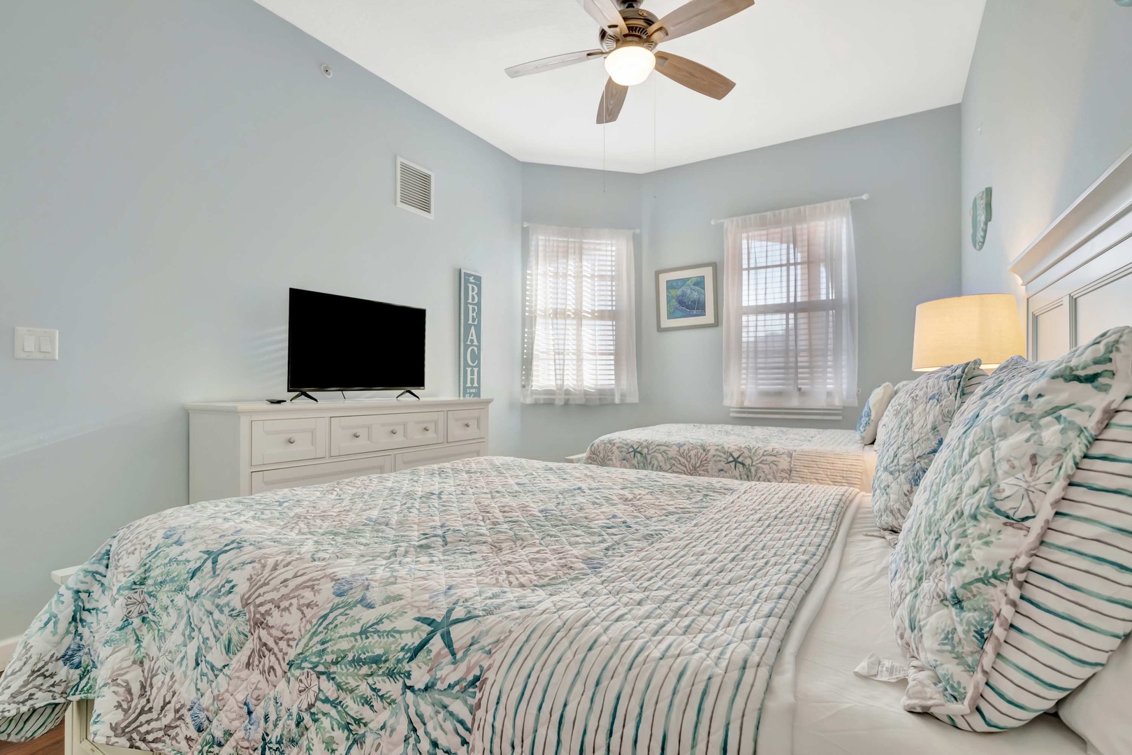 Relax in your serene coastal bedroom with twin beds dressed in calming beach-inspired linens, a ceiling fan to keep you cool, and soft natural light from your windows.