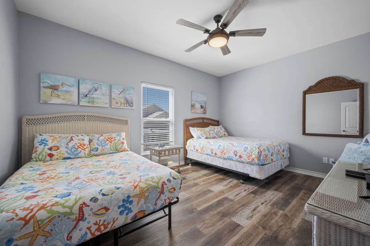 Your coastal guest room features two comfortable beds with vibrant sea-themed linens, a ceiling fan to keep you cool, and cheerful beachside artwork to set the vacation mood.