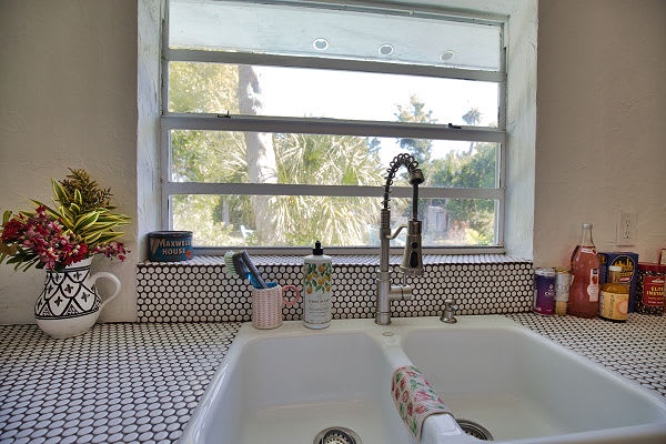 Step into your vacation kitchen where a stylish double sink awaits. The striking geometric backsplash catches your eye while natural light pours through the window, framing palm fronds that sway just outside your retreat.
