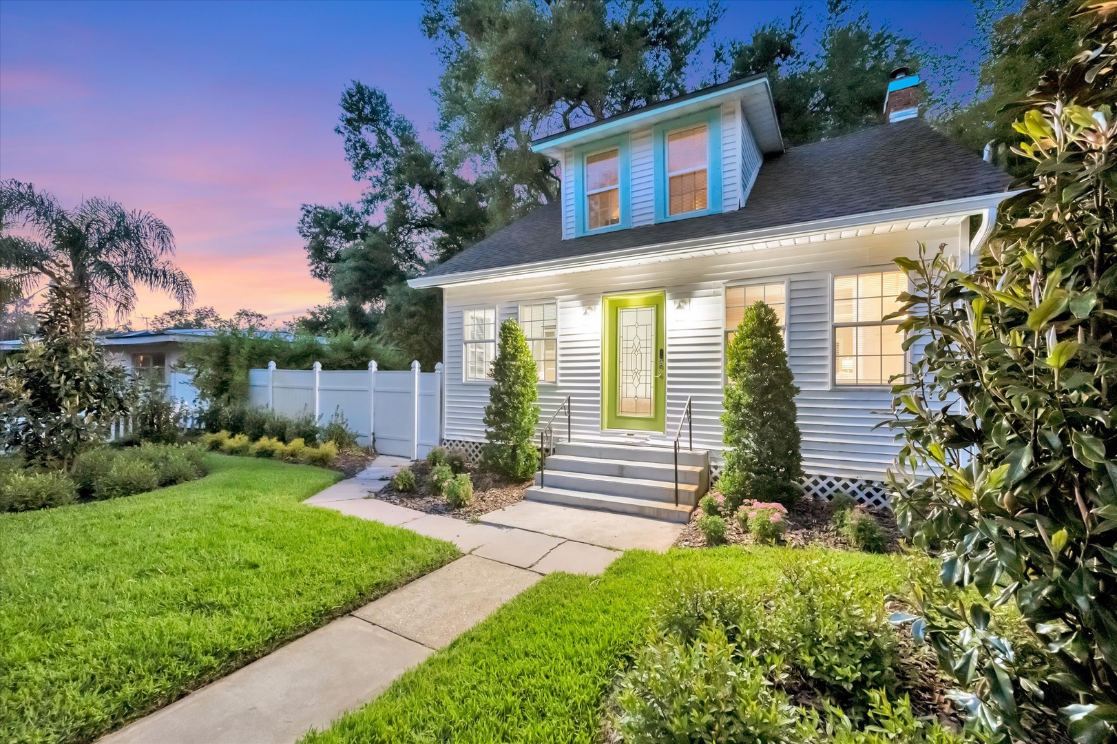 A charming property exterior welcomes guests with its distinctive green entry door, well-manicured lawn, and mature landscaping. The home's turquoise dormer window adds a cheerful pop of color against the warm evening sky.