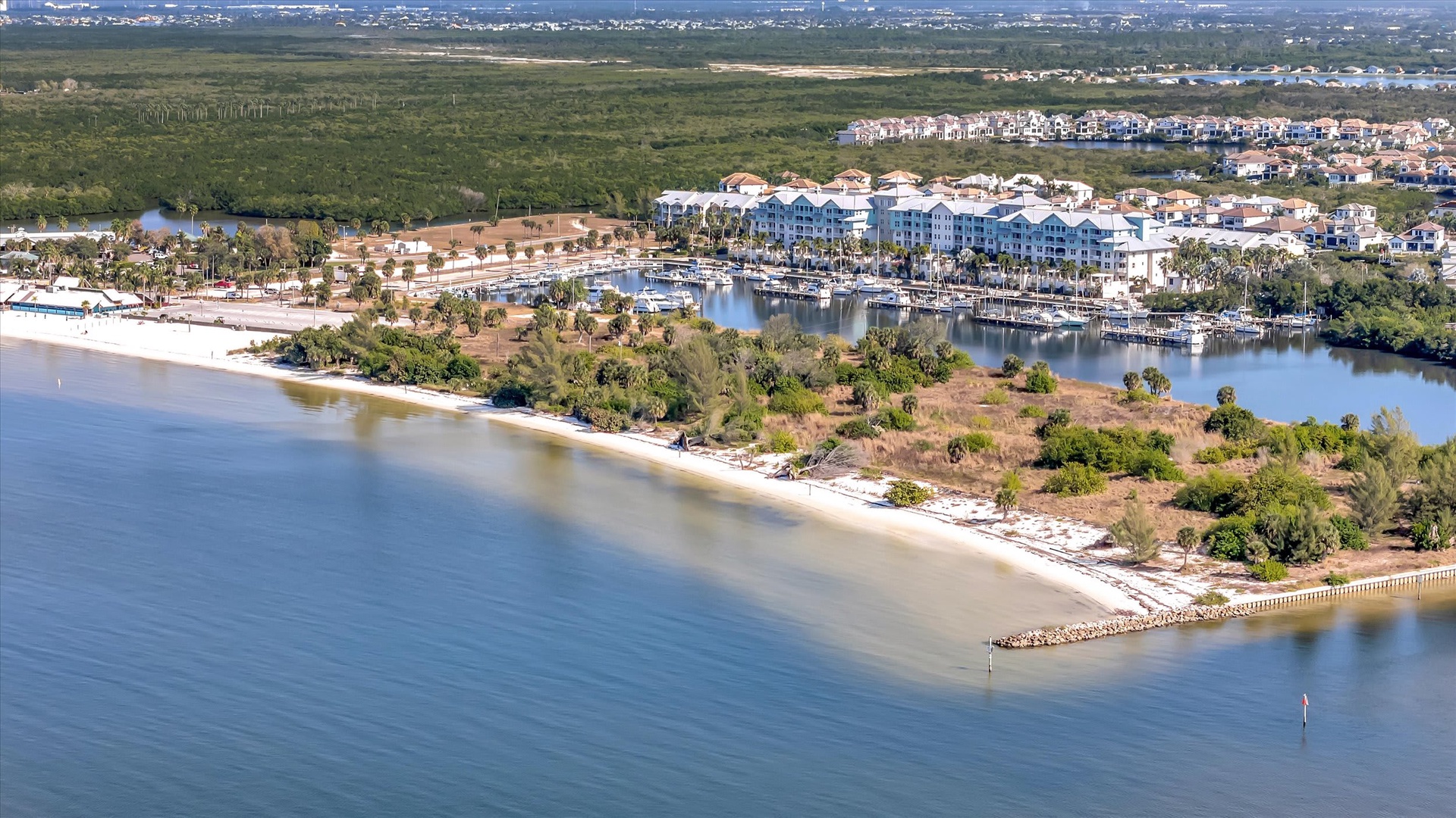 An aerial view showcasing the waterfront resort community, nestled between pristine beaches and a protected marina, with natural coastal landscape and residential neighborhoods surrounding the area.