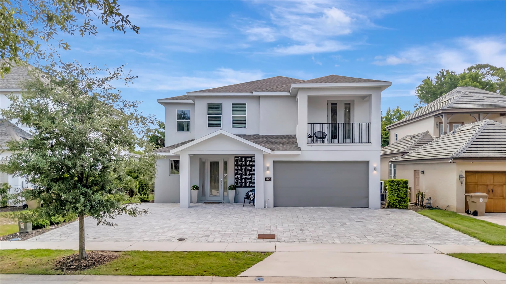 A modern two-story vacation home with a welcoming front entrance and covered balcony, set in a peaceful residential neighborhood with mature trees and landscaped grounds.