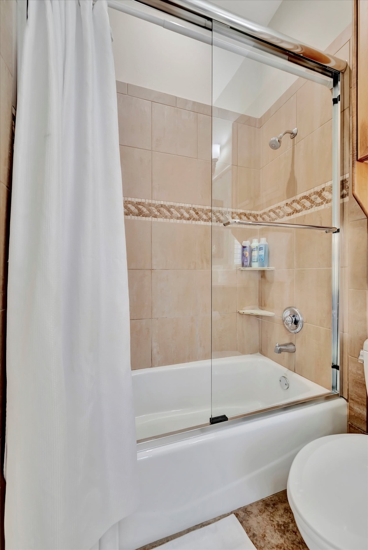 Your private bathroom features warm beige tile with decorative accents, a tub-shower combo with glass doors, and fresh towels ready for your arrival.