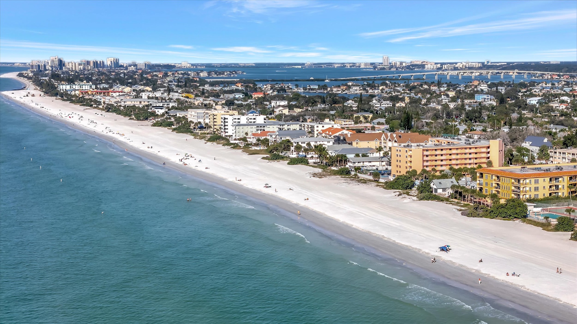 An aerial perspective showcasing the beachfront neighborhood where the property is located, with pristine white sands meeting turquoise waters and coastal buildings lining the shore.