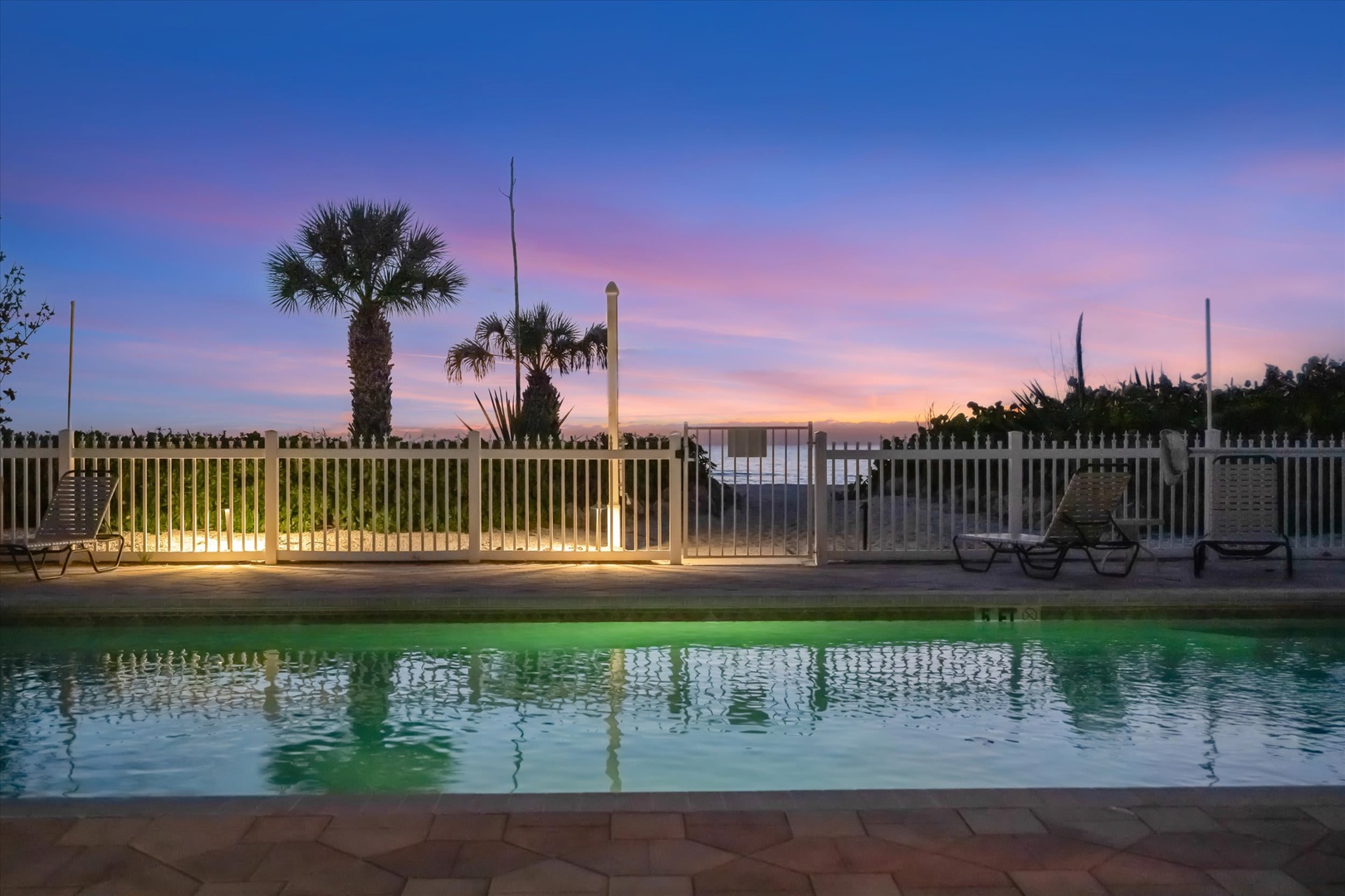 The pool glows softly as the sky melts into pastel pinks and oranges at twilight, with palm trees silhouetted beyond the fence—a peaceful moment to end your day.