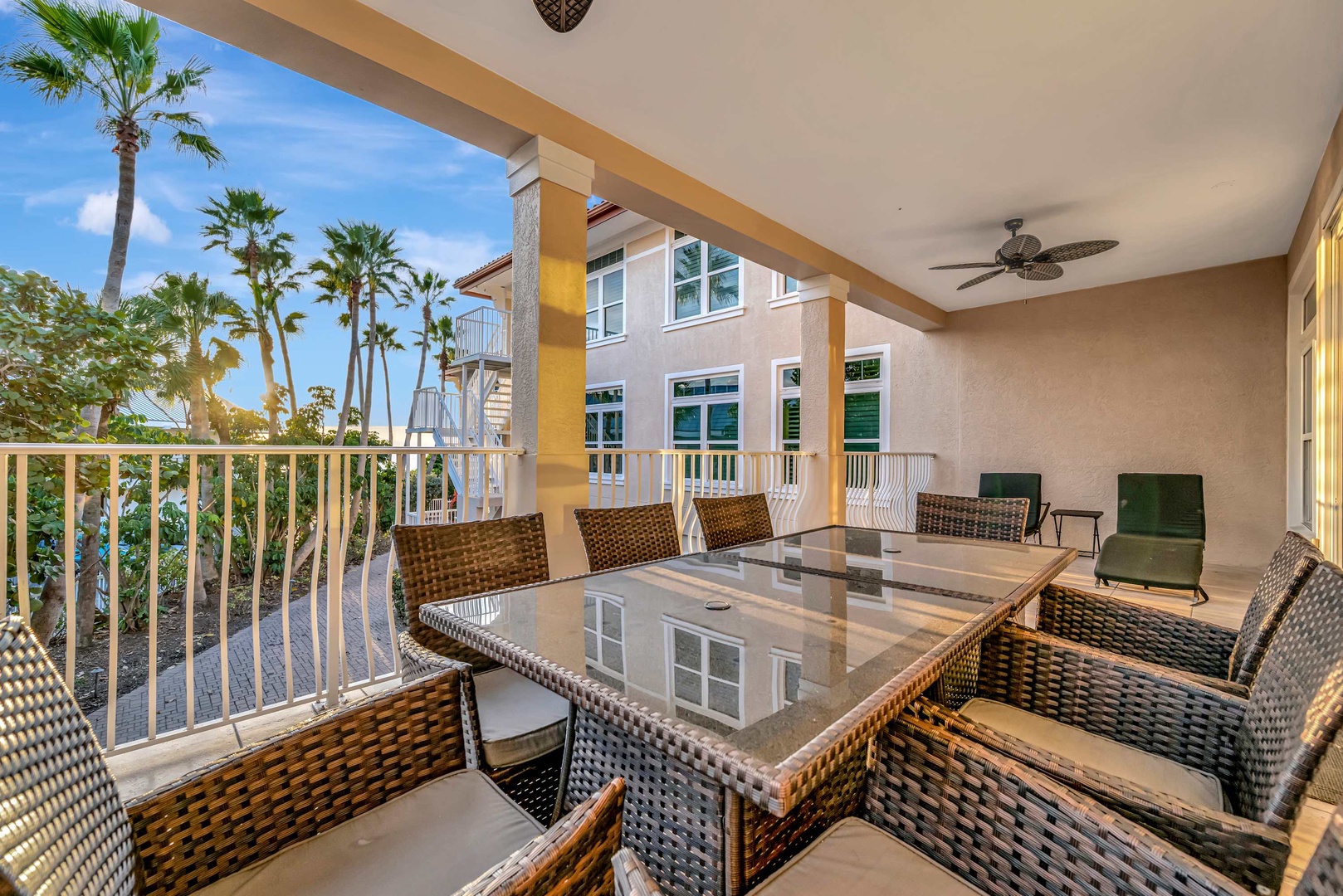 Spacious covered balcony featuring a large dining table and comfortable woven seating—perfect for gathering under ceiling fans while enjoying tropical breezes and palm tree views.