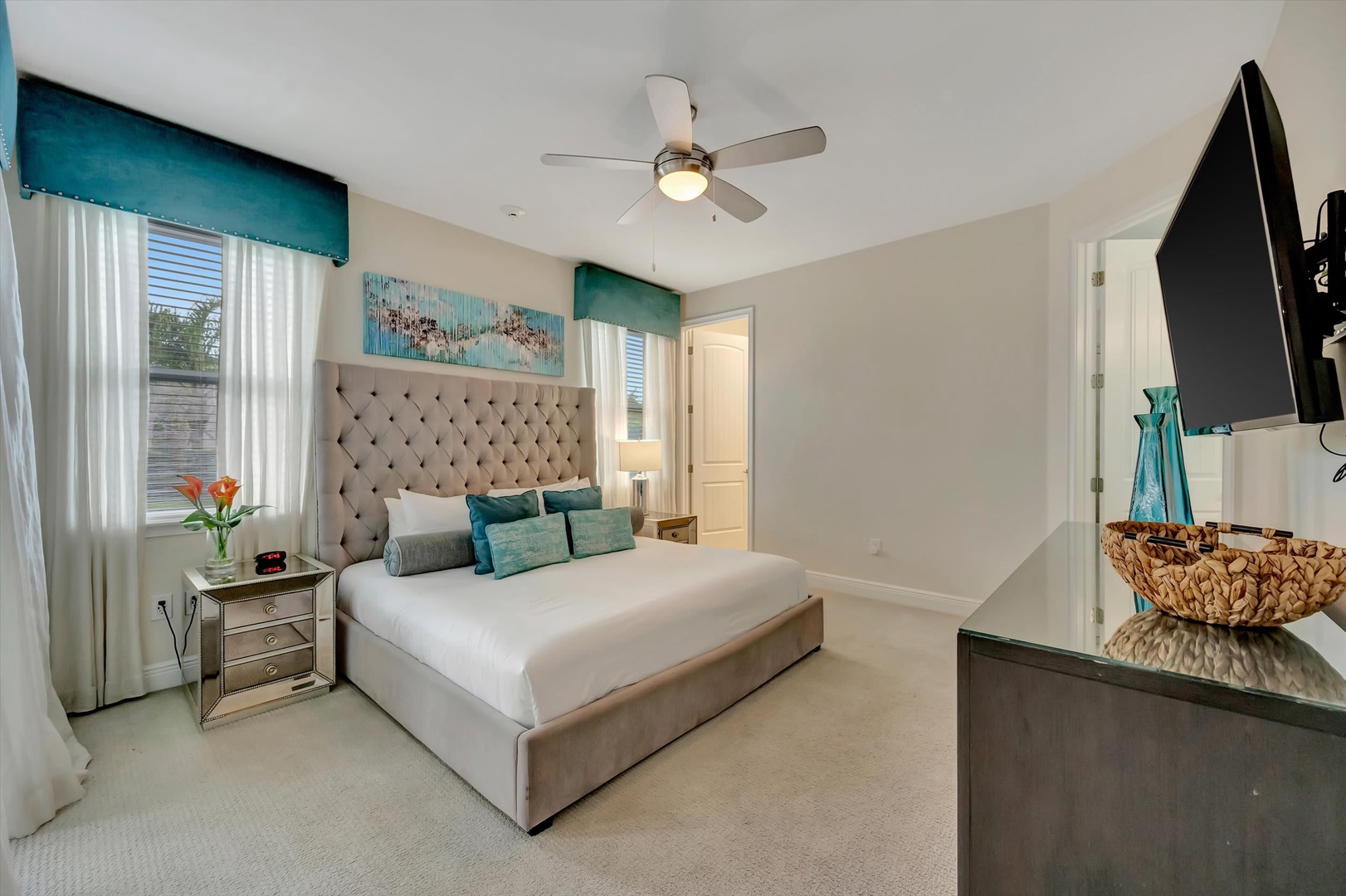 Your serene bedroom retreat features a plush upholstered bed, crisp white linens with turquoise accents, and gentle natural light filtering through elegant curtains. The ceiling fan keeps you comfortable while you unwind with a favorite show.