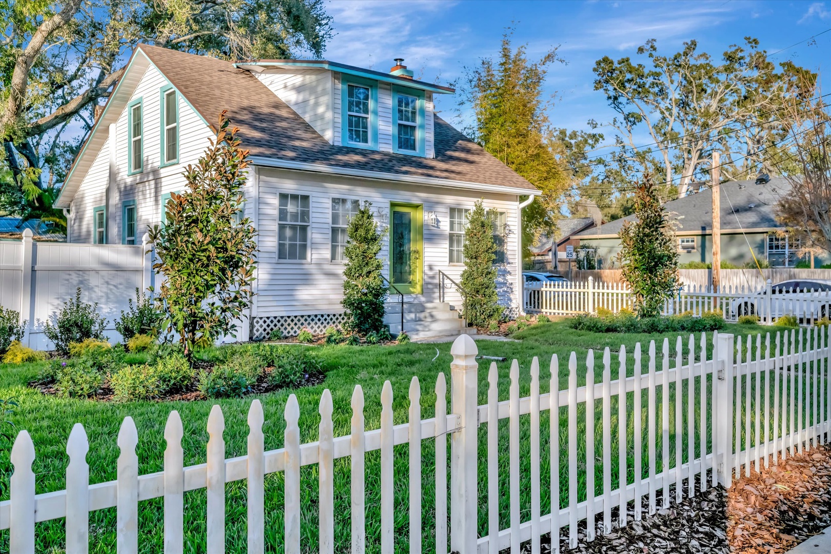 Charming white cottage with a welcoming green door and bright shutters, nestled in a peaceful neighborhood setting with mature trees and picket fence.