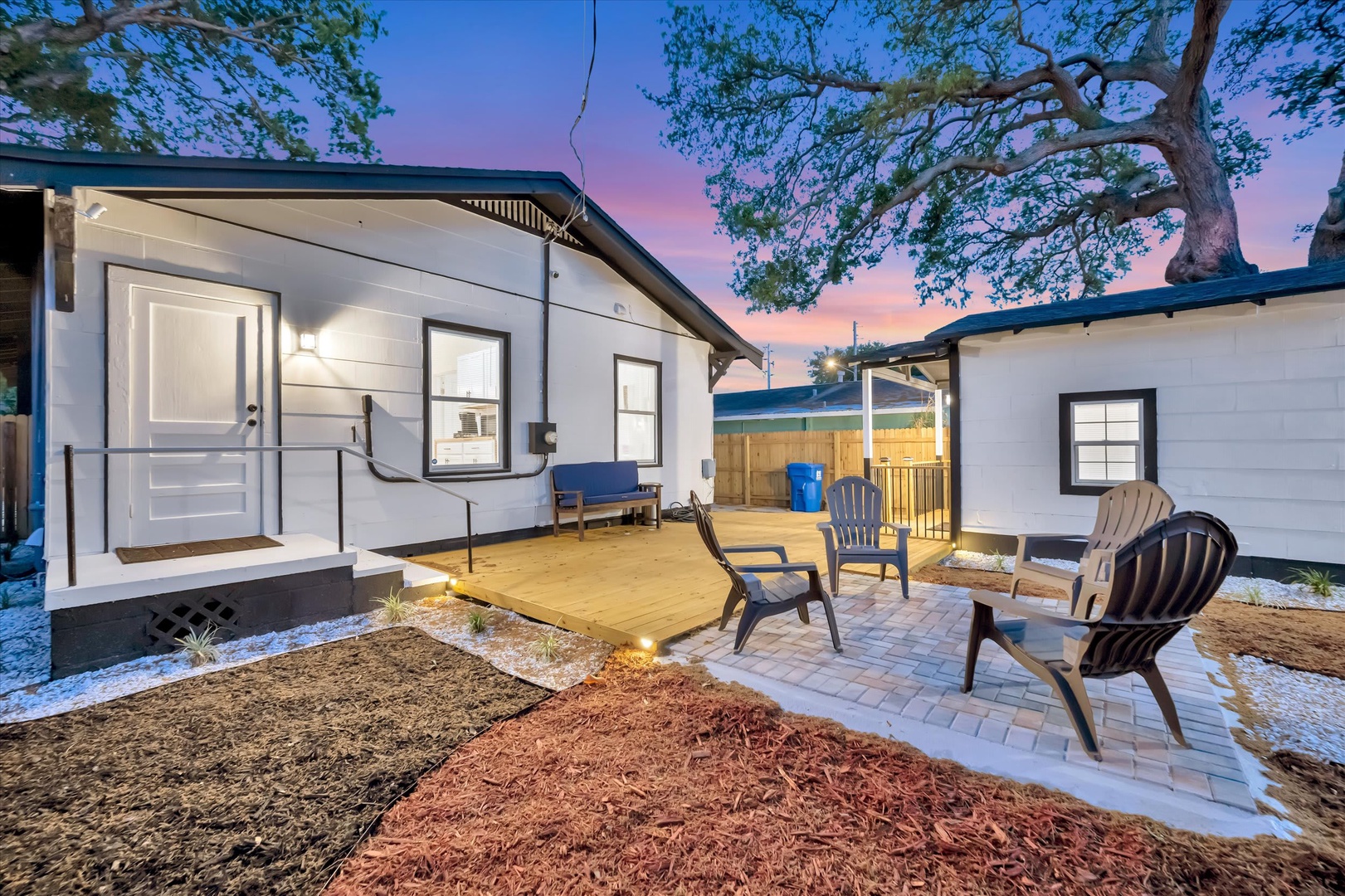 A modern cottage nestled beneath mature trees, featuring a welcoming deck and patio with Adirondack chairs perfect for evening relaxation. The property's crisp exterior and warm lighting create an inviting arrival experience as dusk settles in.
