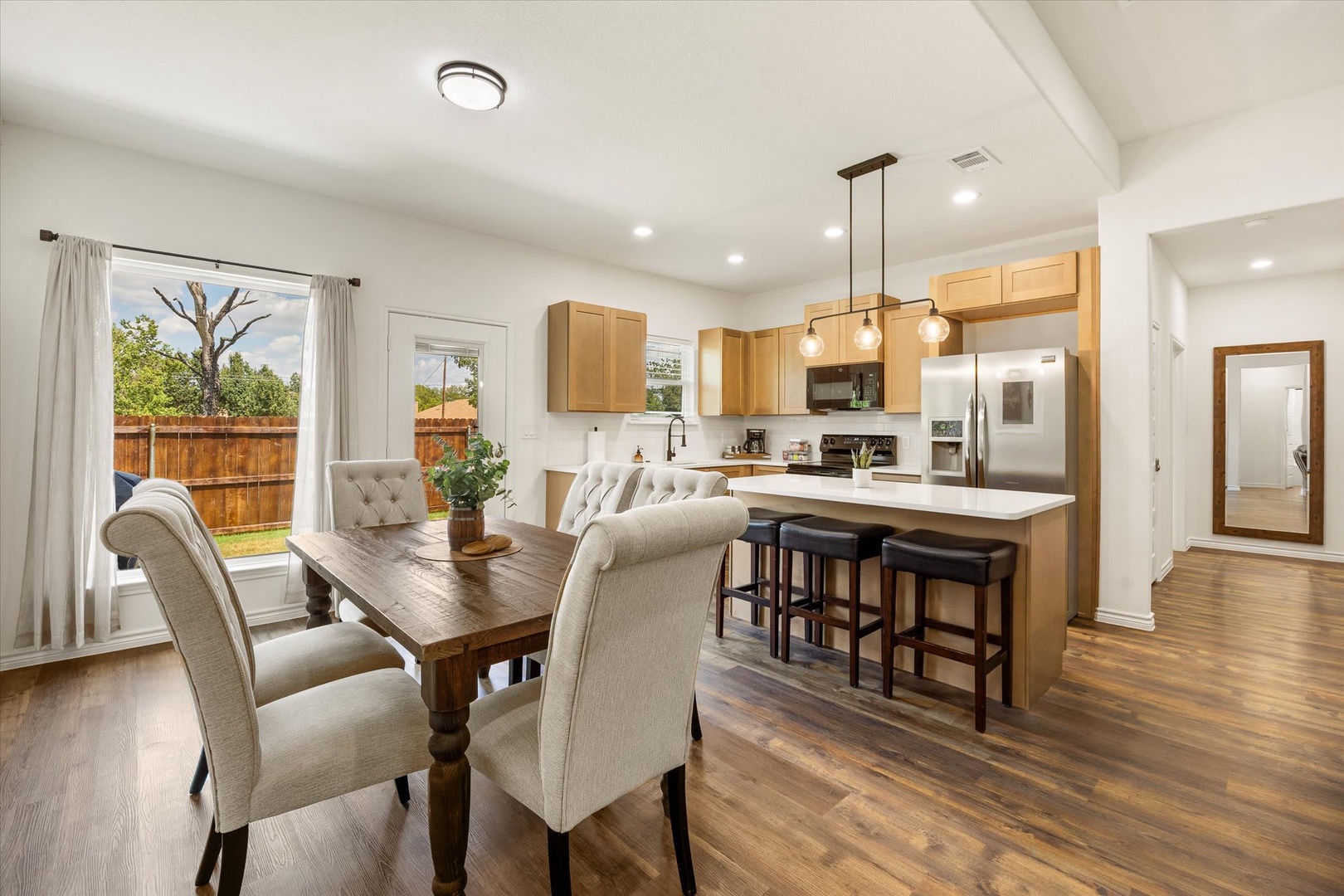 Open-concept kitchen and dining area with warm wood tones, modern finishes, and a central island. Natural light streams through large windows overlooking the private backyard.
