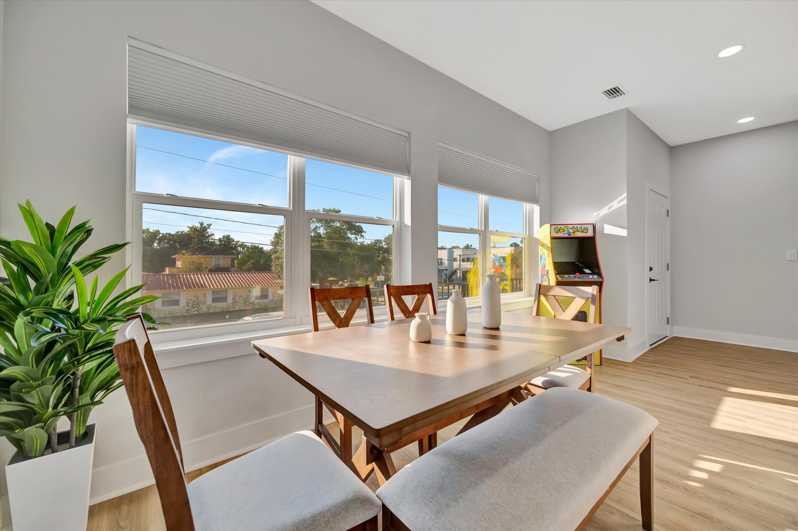 Gather around the dining table with sunlight streaming through large windows, blending comfort and playful vibes with an arcade game ready for your downtime fun.